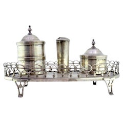 Silver inkwell w/ two tanks, resting on a base w/ a grate 
four cut-out feet