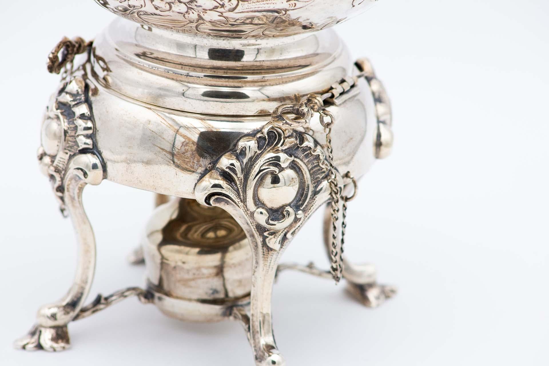 Description : Bouilloire en argent à motifs floraux repoussés, éléments végétaux, bouton en forme de rose sur le couvercle, reposant sur un dessous de plat à pieds surélevés en forme de feuilles d'acanthe et une lampe. Métal : Argent. Origine :