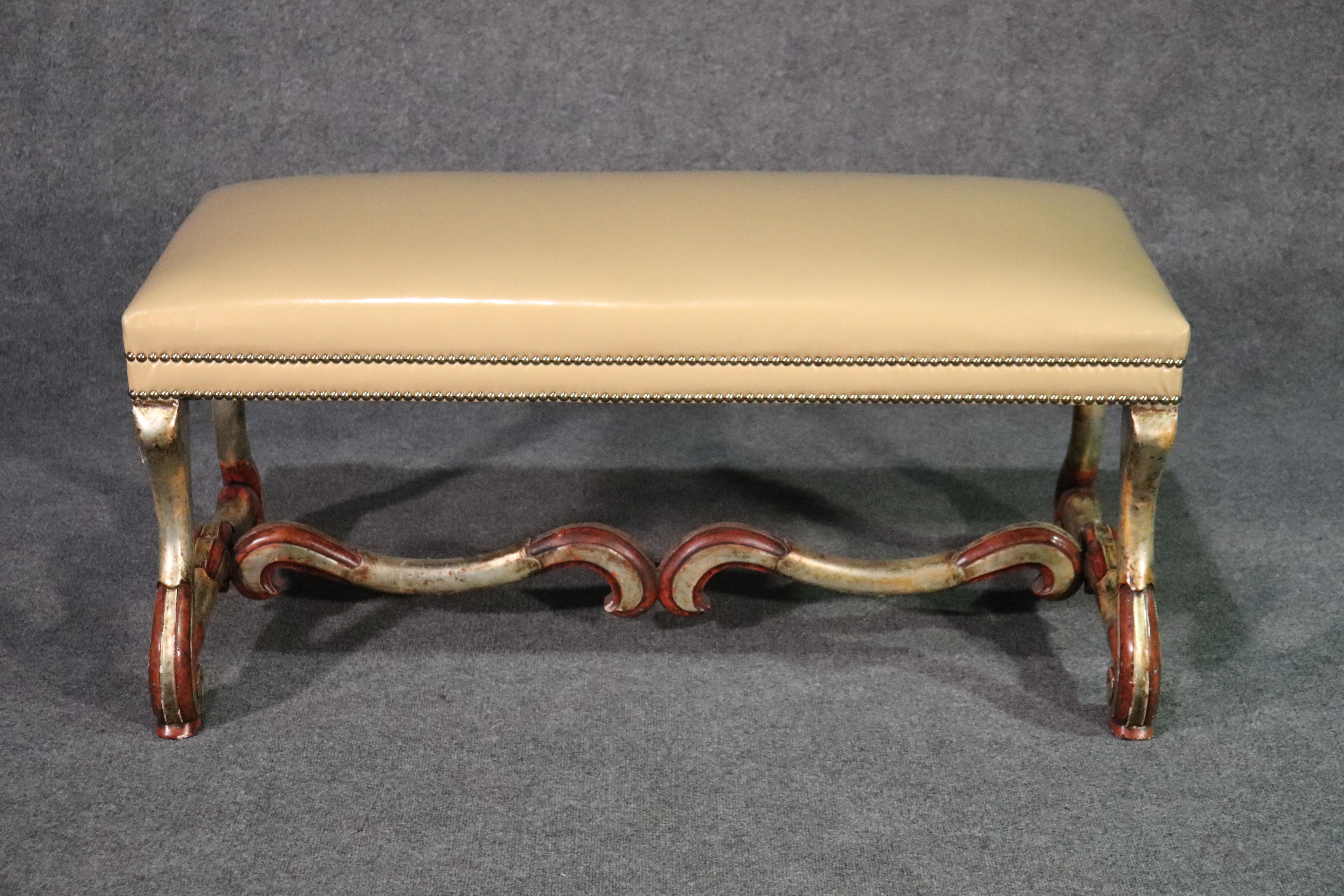 This is a beautiful window bench done in silver leaf over a red bole ground. The styling is classic French mutton legs and the upholstery is a tan patent leather or vinyl. The upholstery as well as the frame are in good condition. The piece dates to