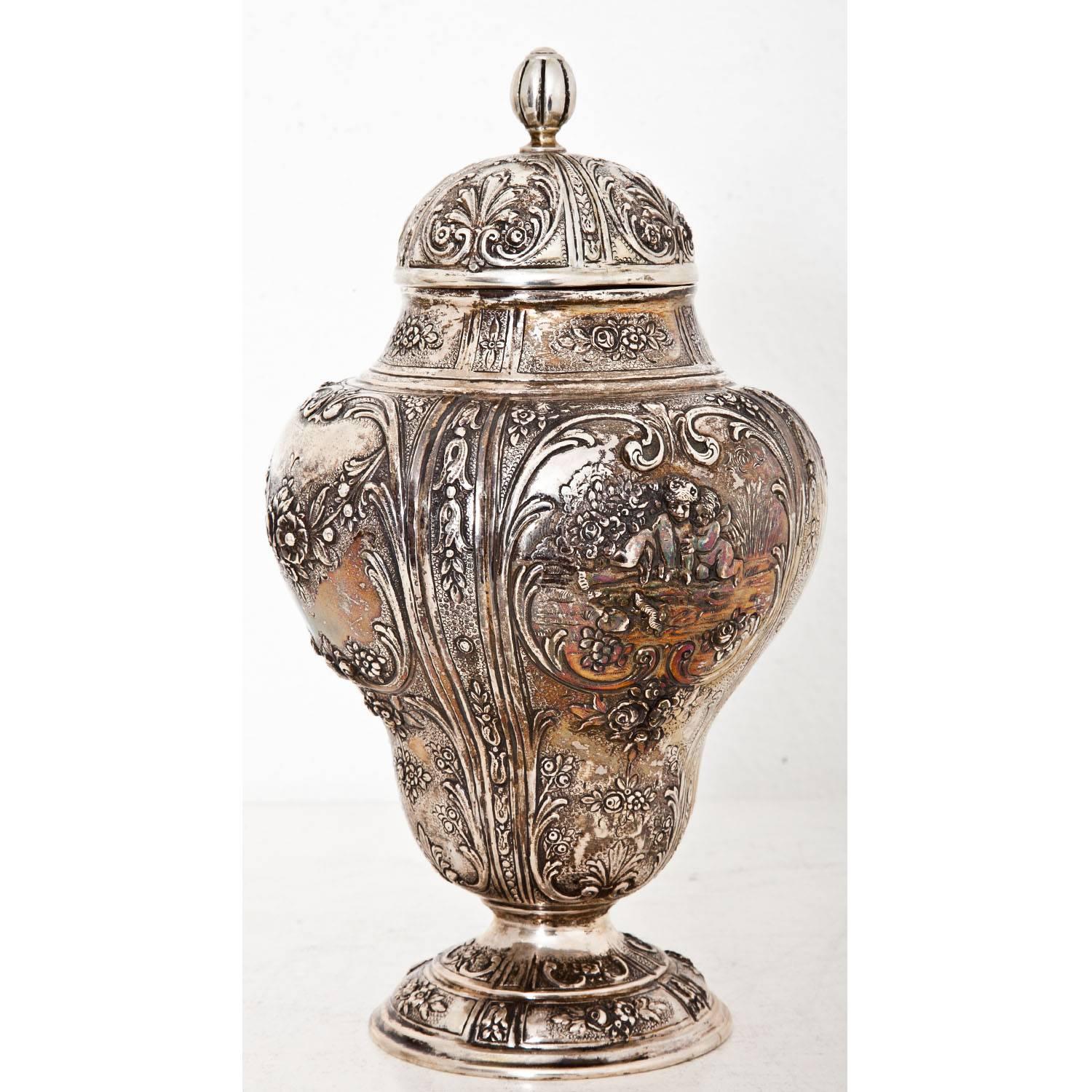 Silver lidded vase on a round stand with a bellied body. The wall is decorated with roses and leaves and shows cupids in cartouches. The urn is stamped at the bottom for Germany, silver 800 and maker’s mark OS.