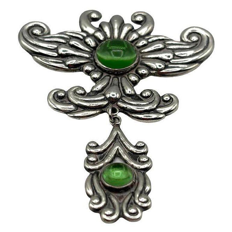 Diese Jugendstil-Brosche aus Silber mit ihrem atemberaubenden Mittelstück aus grünem Achat verkörpert die fließende, organische Schönheit der Epoche. Zarte Silberarbeiten umgeben den Stein und formen verschlungene Blumen- und Rankenmotive, die die