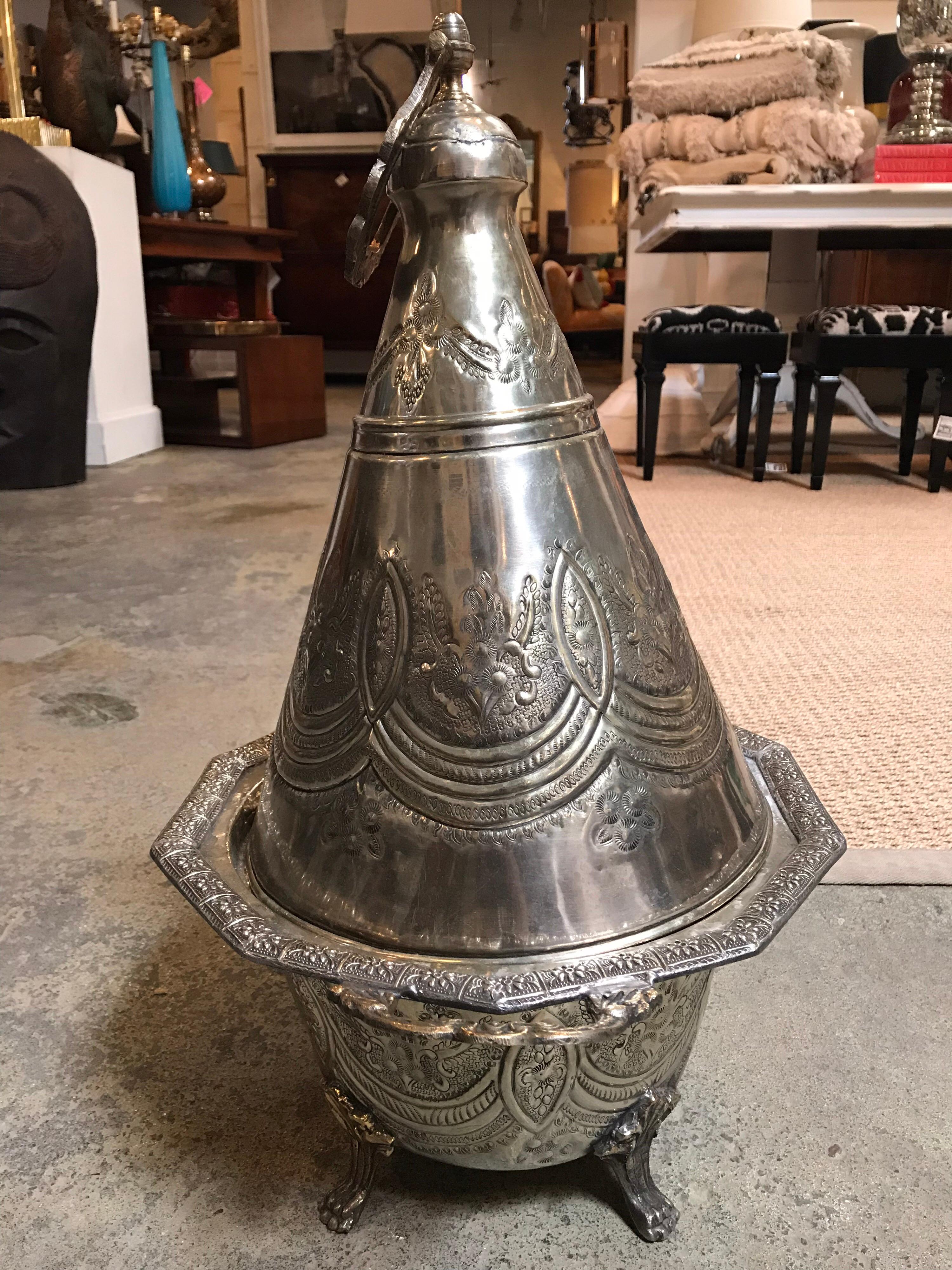 This silver metal Moroccan tajine is a tall representation of exquisite Moroccan detail. It was made in a Repousse style and has handles on two if it's sides with four clawfoot feet to support it. There is also a handle atop the lid that is freely