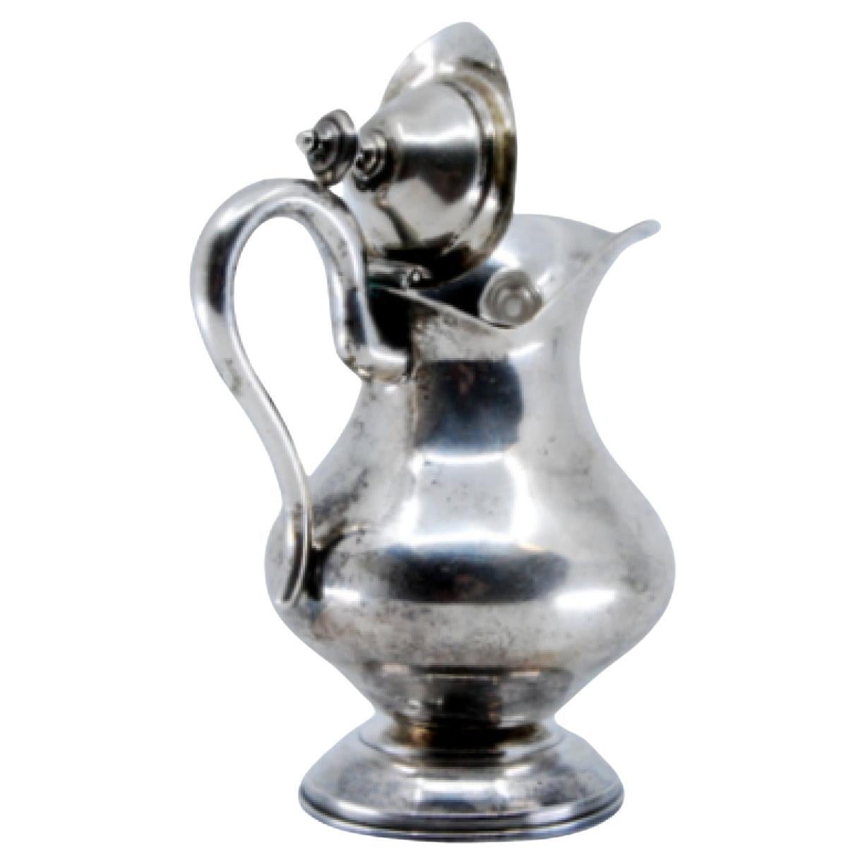 Silver milk jug w/ simple handle, lid w/ knob & resting on a base w/ frieze