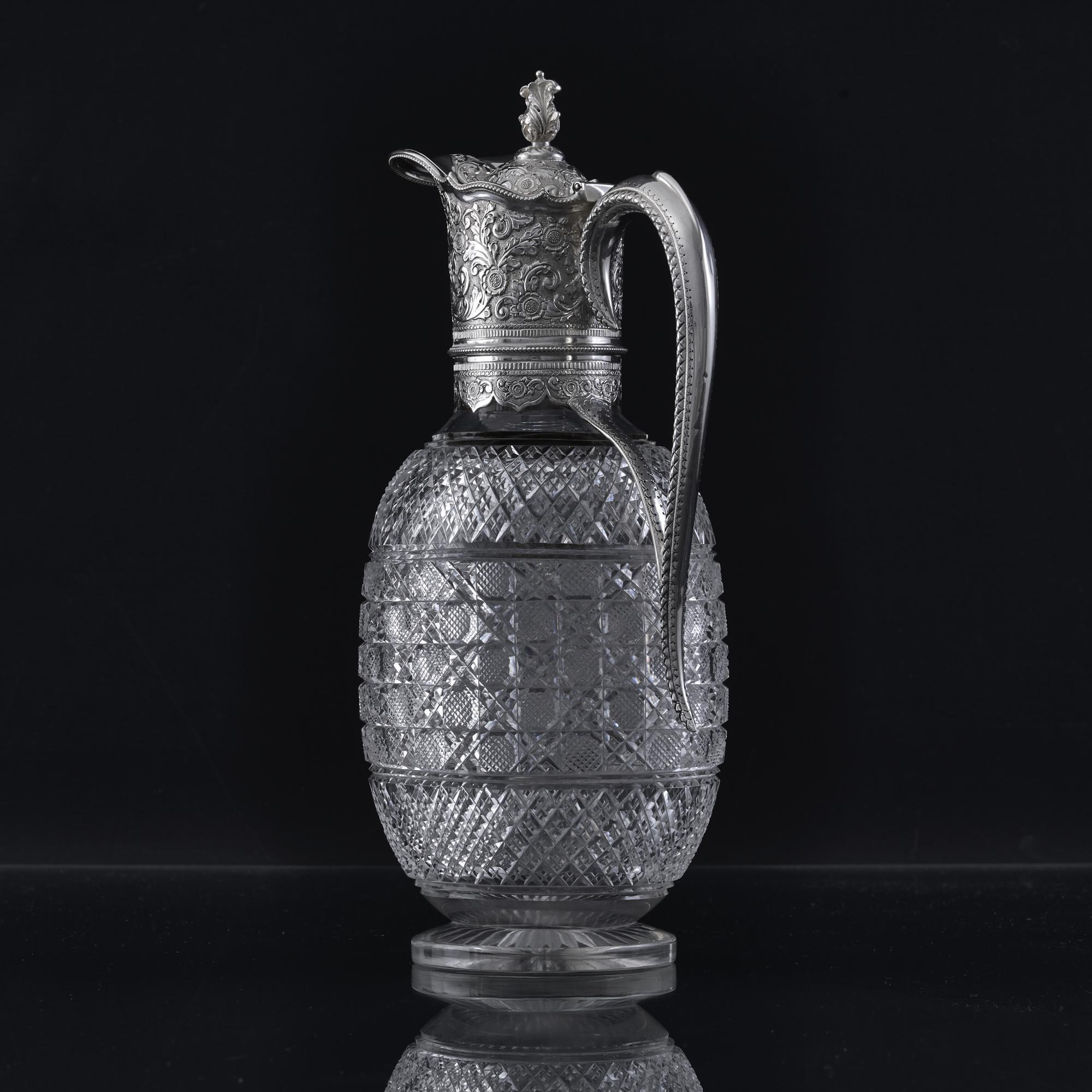 This antique Victorian wine jug has a round cut crystal body featuring three bands of lattice cut detail separated by sections of subtle leaf and flower decoration. The silver collar is hand-chased with bas-relief decoration of scrolling foliage