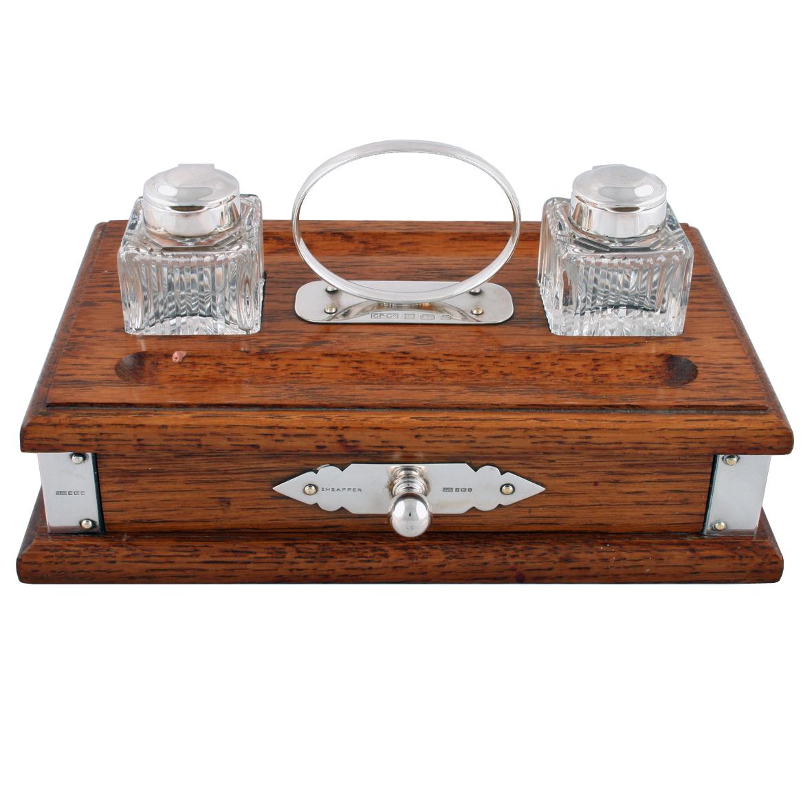 A late 20th century oak inkwell and pen stand.

The oblong stand has a single drawer, compartments to sit two inkwells, with sterling silver hinged lids, two pens and has a hall marked sterling silver drawer handle, stand handle and corner