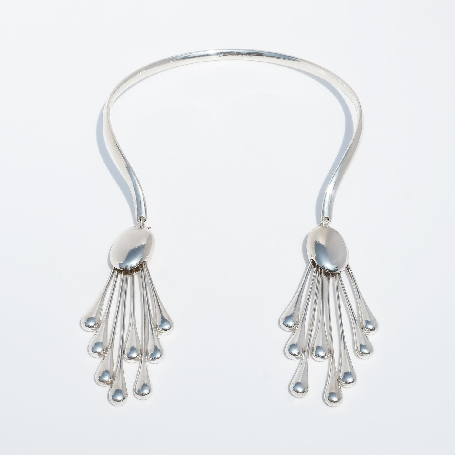 This sculptural silver neck ring features an open, curved form that rests elegantly around the neck. At each end hangs a detachable pendant composed of a smooth, oval element—resembling a polished shield—from which eight elongated, drop-shaped rods