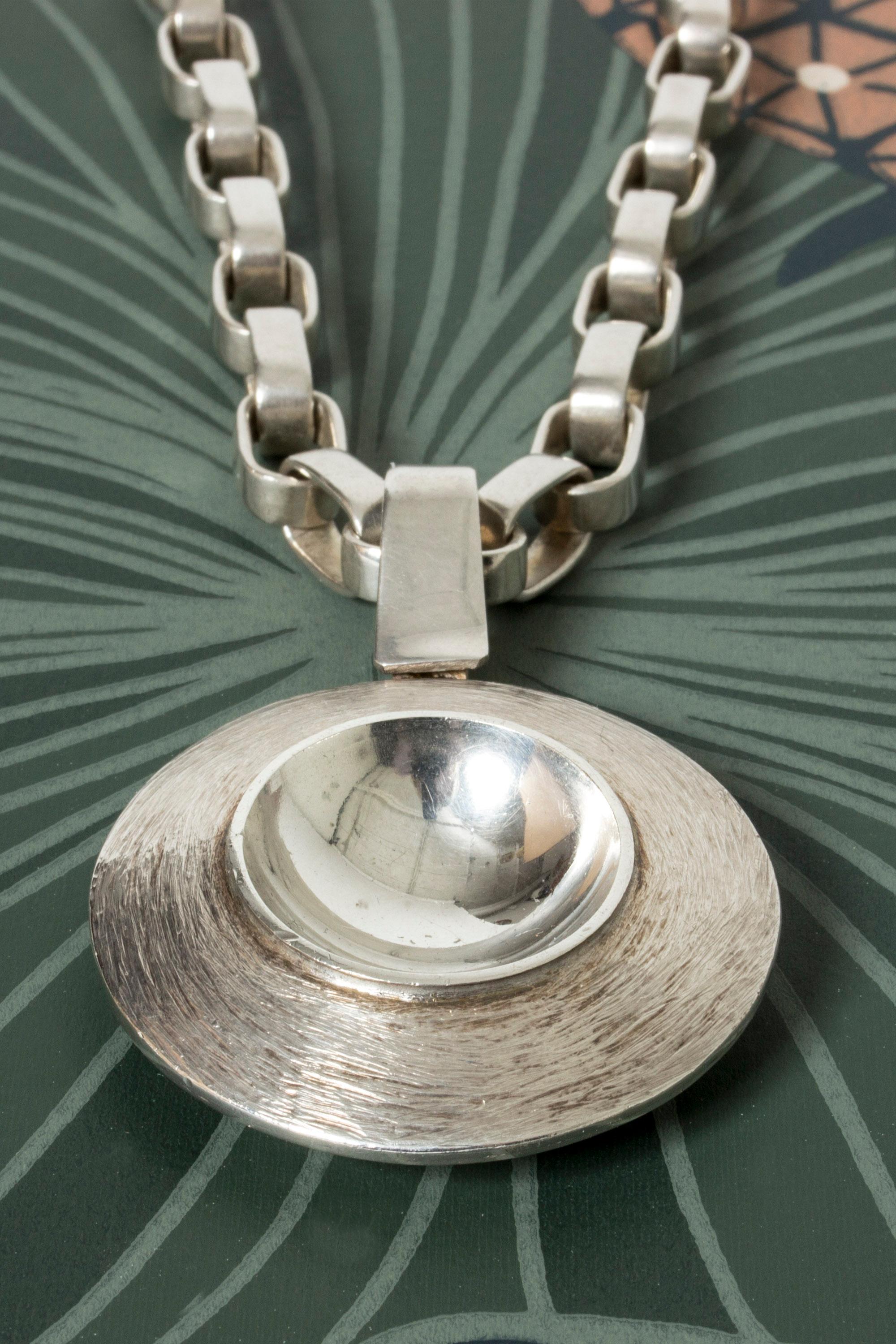 Magnificent silver necklace by Sven-Erik Högberg with a hypnotizing, round pendant on a thick chain that can be worn just as well on its own. Wonderful design and craftsmanship.

Year: 1974 (pendant) and 1978 (chain)
Dimensions: Height 6.5 cm