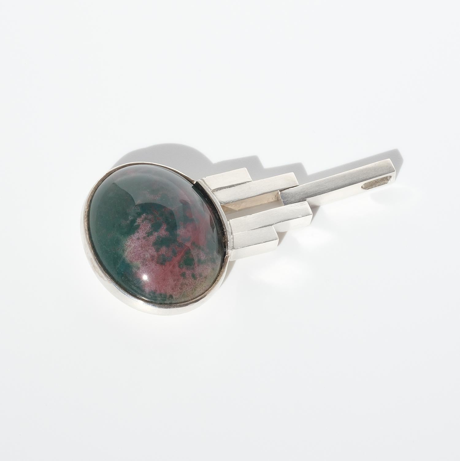This silver neck-ring has a pendant made out of sterling silver and a cabochon cut variegated jasper stone in dark green and red. The top of the pendant is decorated with a spiral made out of five silver bars, a creation that might bring your
