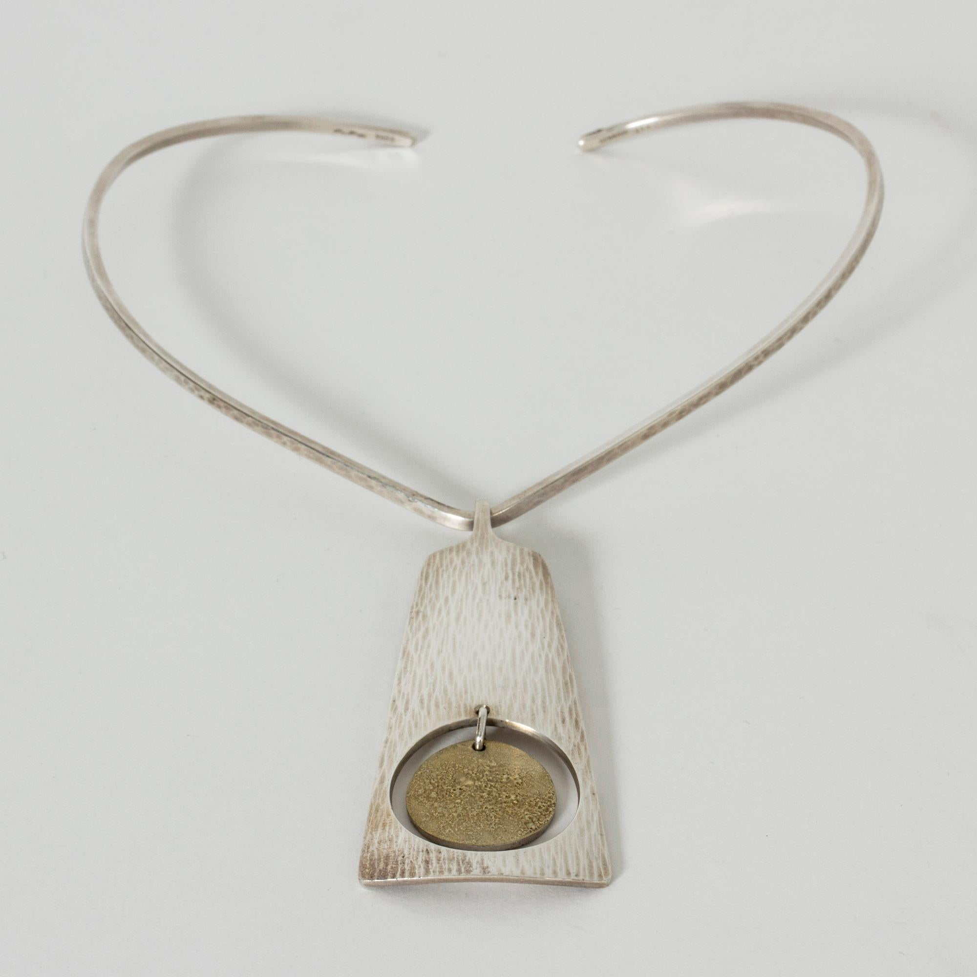 Striking silver neckring by Bent Gabrielsen Pedersen with a long pendant with a hand-hammered surface. In the cut out sphere in the middle of the pendant hangs a gilded disk with an acid treated surface that gives it a foaming look. Great contrasts,