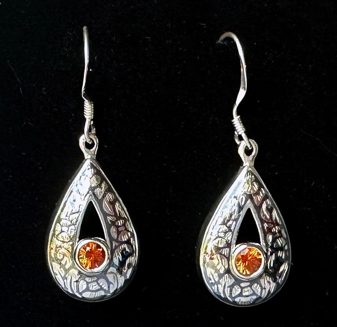 Silver Nielloware Earrings set with Orange Sapphires. 

Nielloware is a Thai Silver craft that is practiced in the southern provinces of Thailand. Using a blackened Nickel Alloy to inlay a cultural design into the Silver, the alloy provides a stark
