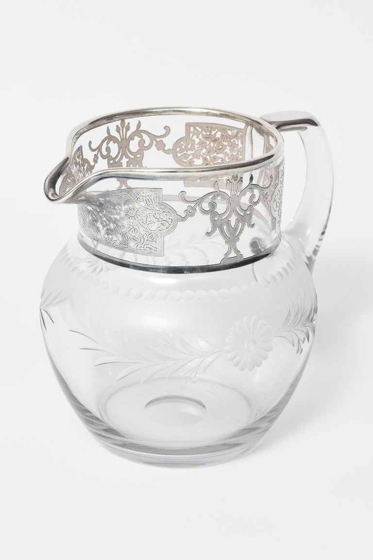 Clear glass water or lemonade pitcher decorated with etched daisy flowers with leaves and dots on each side. The top 2.5” section has silver overlay featuring leaves and urns with flower sprays. The pitcher has a handle and the thumb area has silver