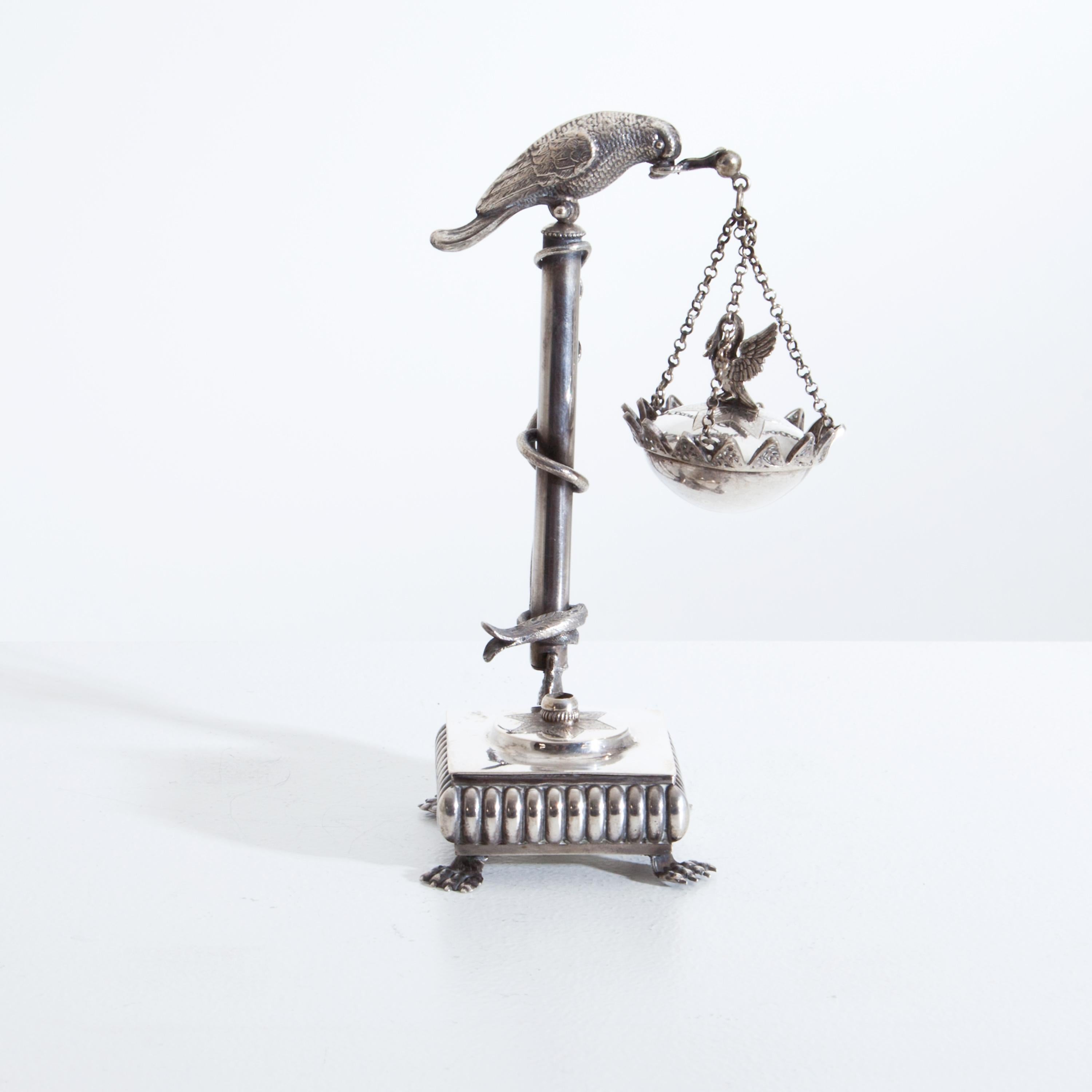 A square oil lamp standing on claw feet with a straight column surrounded by a snake and crowned by a bird. Hanging down from it is the bowl with lid and swan knob.