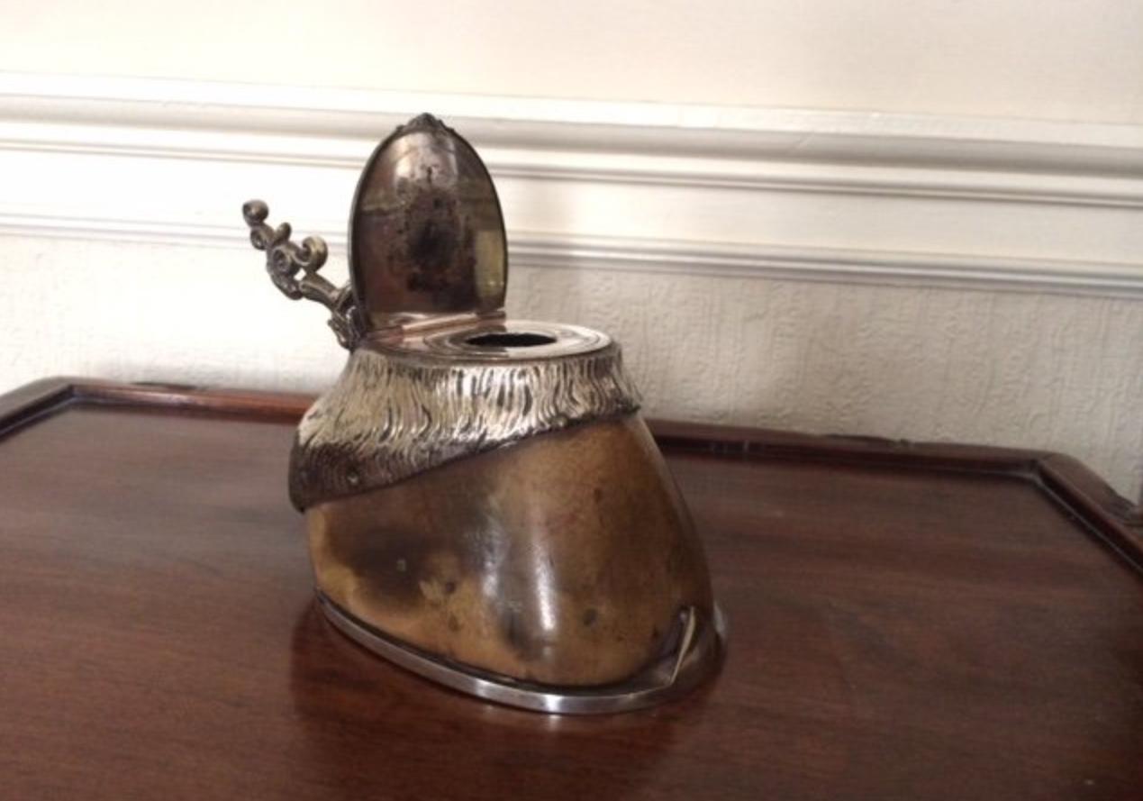 A late 19th century silver plate mounted horse hoof inkwell, the lid and horseshoe of silver plate, the hinged lid opening to the glass inkwell with silver plate collar, with a decorative pen rest above, the the hoof with good feathered detail