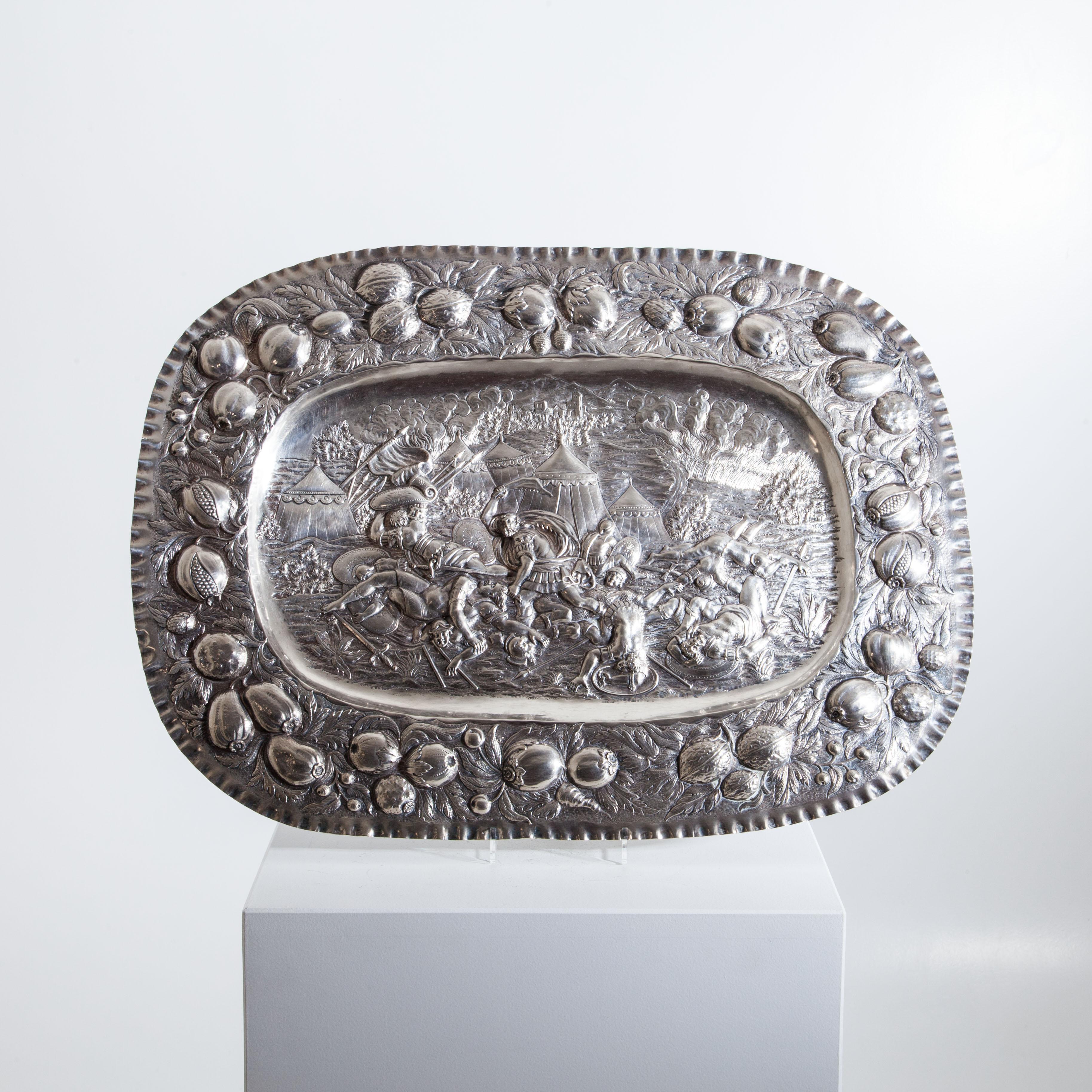 Oval silver plate with embossed nut and fruit decoration on the rim and detailed, dramatic battle scene in the mirror, probably depicting Samson slaying a Philistine.
    