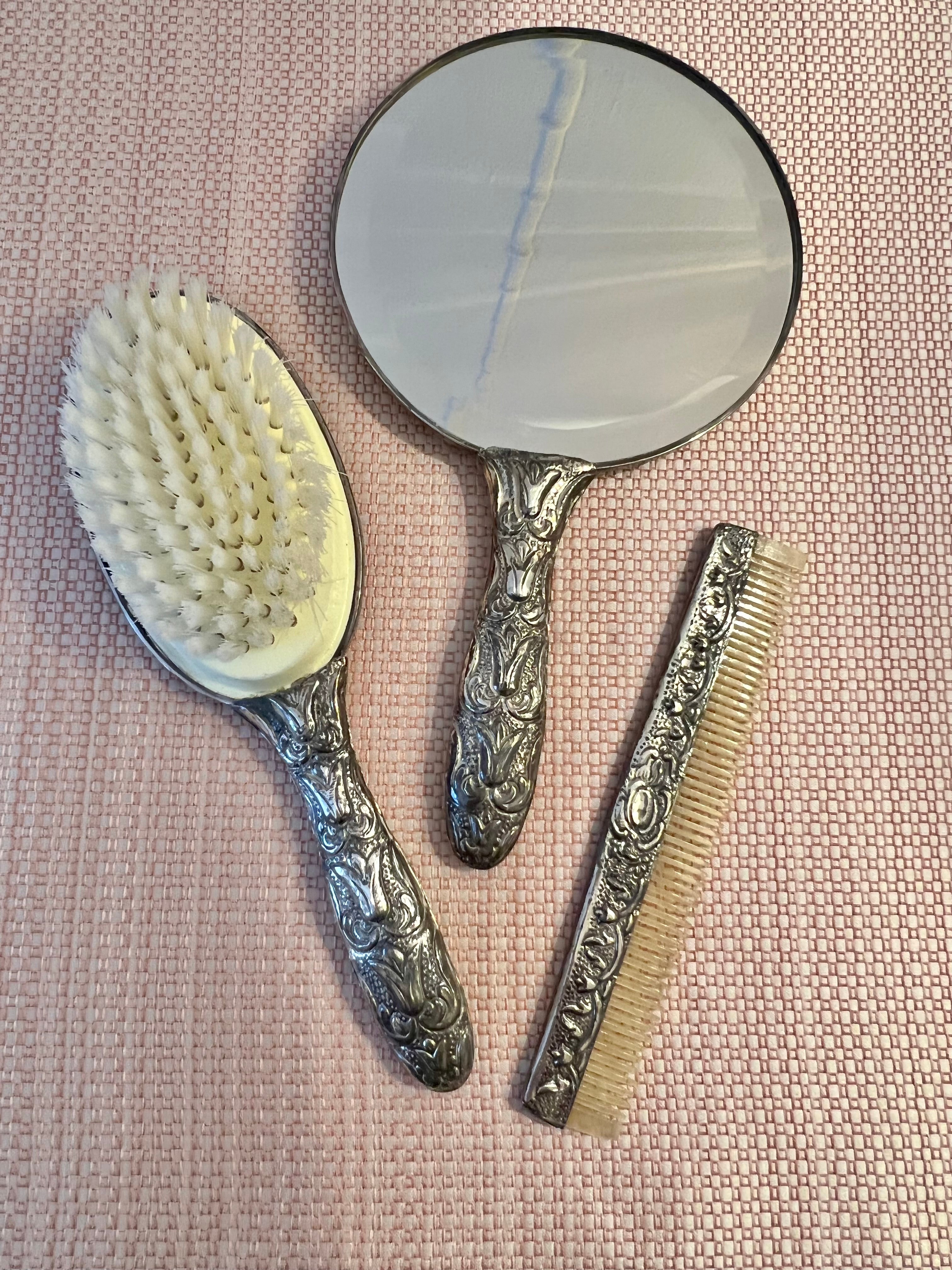 Vintage, never used, vanity set with a bristle brush, comb and mirror. Each piece fits neatly into a velvet carrying case with hinge closure. 

Each piece features an ornate silver plate repousse brocade. Brush and mirror have been engraved with