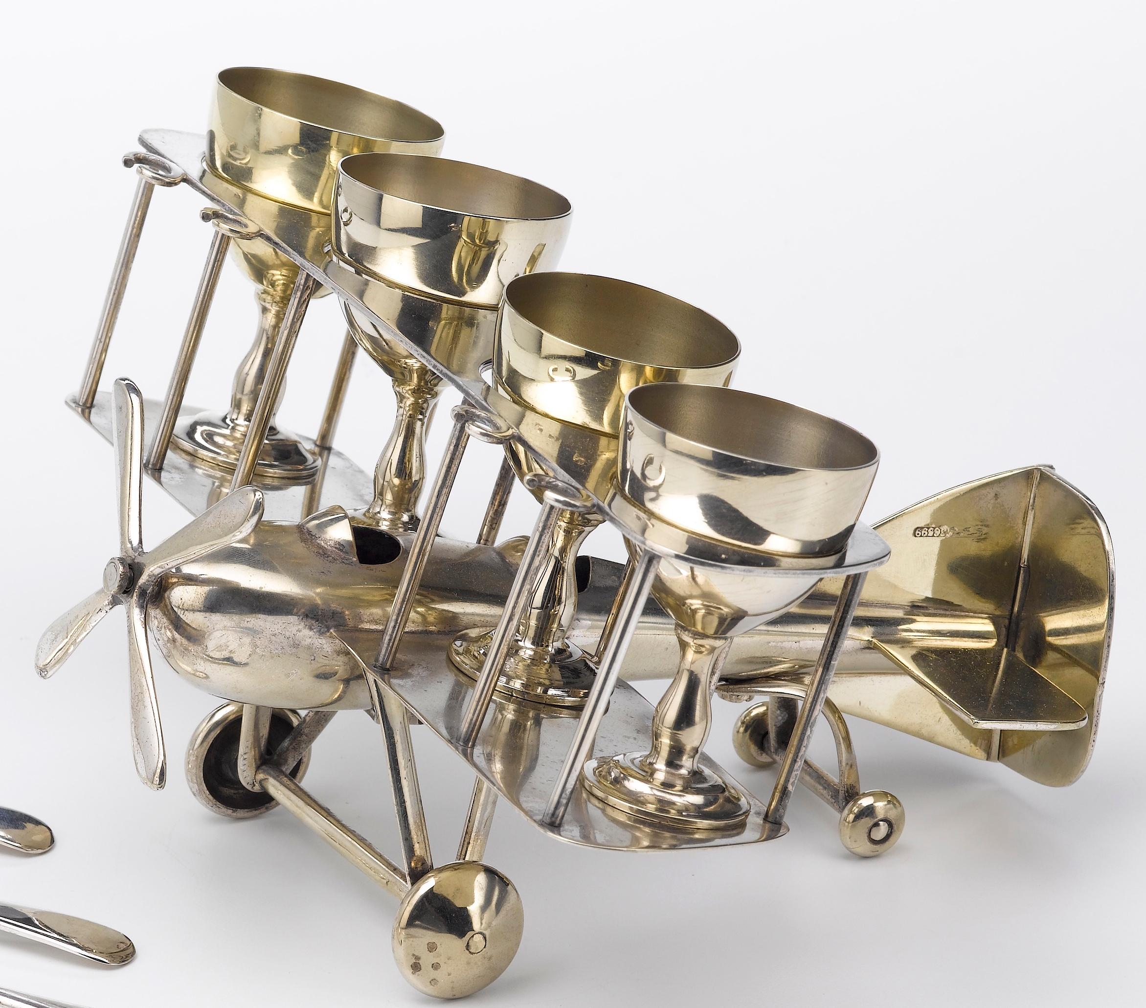 This is a beautiful silver-plated egg cup and spoon set with an airplane motif, dating to the early 1910s. The set includes four matching footed egg cups. The cups sit upon the stacked wings of a biplane. Four small egg spoons accompany the set. The