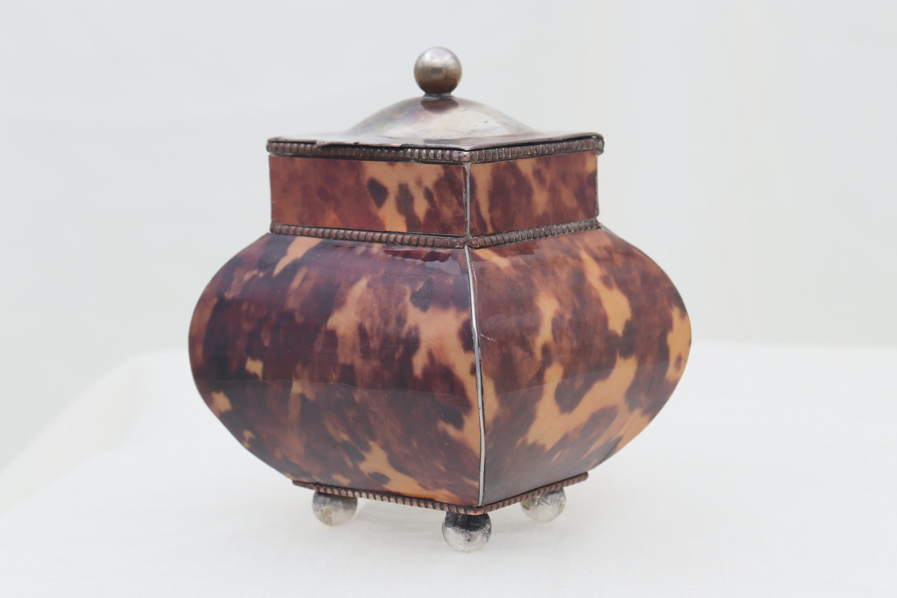 The bombe shape of this small tea canister is made of silver plate and covered with faux tortoiseshell. The domed lid is topped with a ball knop and the piece sits on four silver plated ball feet. There are three gadrooned edges and the panels of