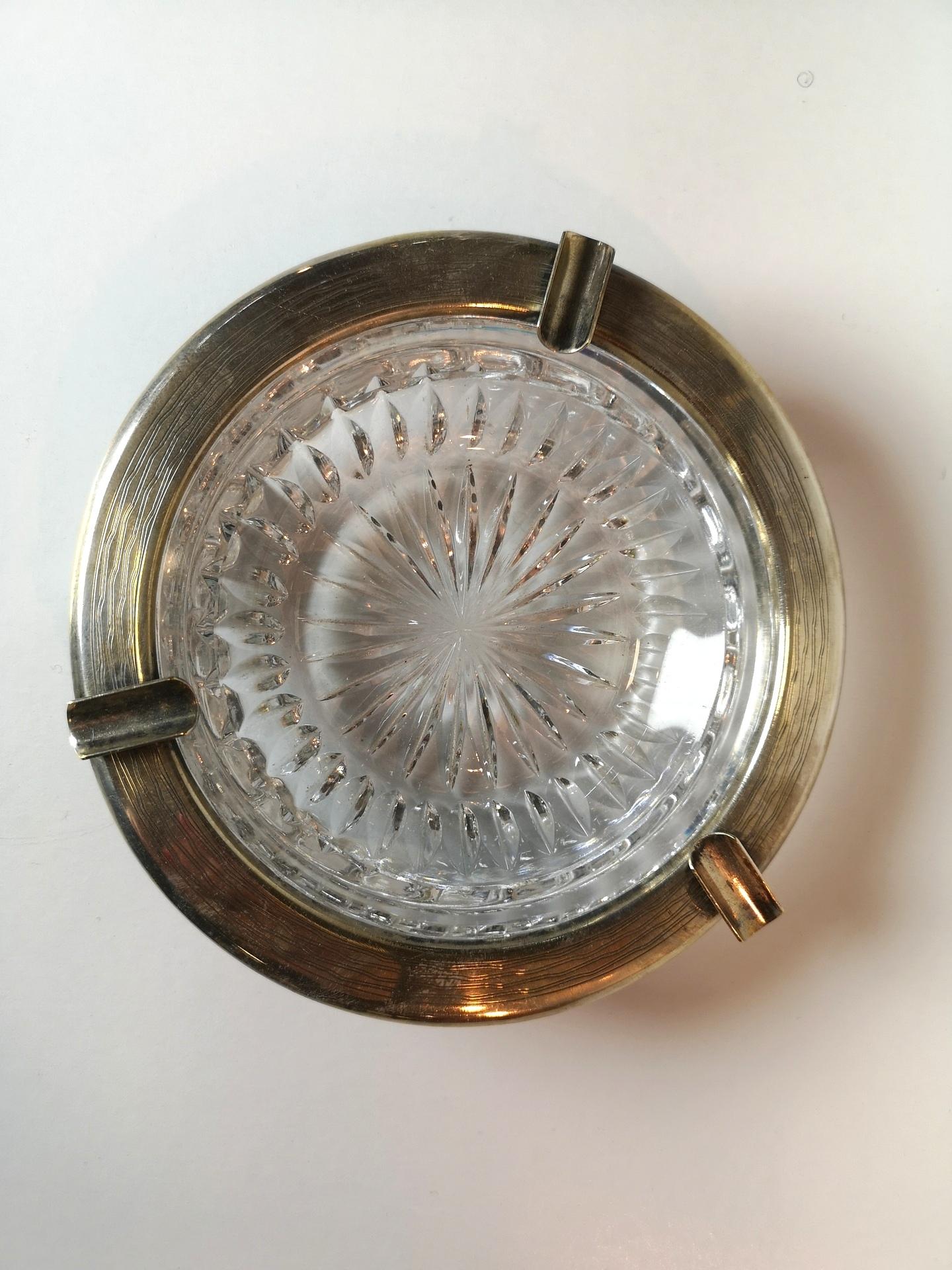 This chiseled crystal glass Art Deco ashtray features a silver plated brass rim, parts of which extend to the bottom of the object, forming a tripod stand. This combination of metal and crystal glass creates a stylish and elegant table top design