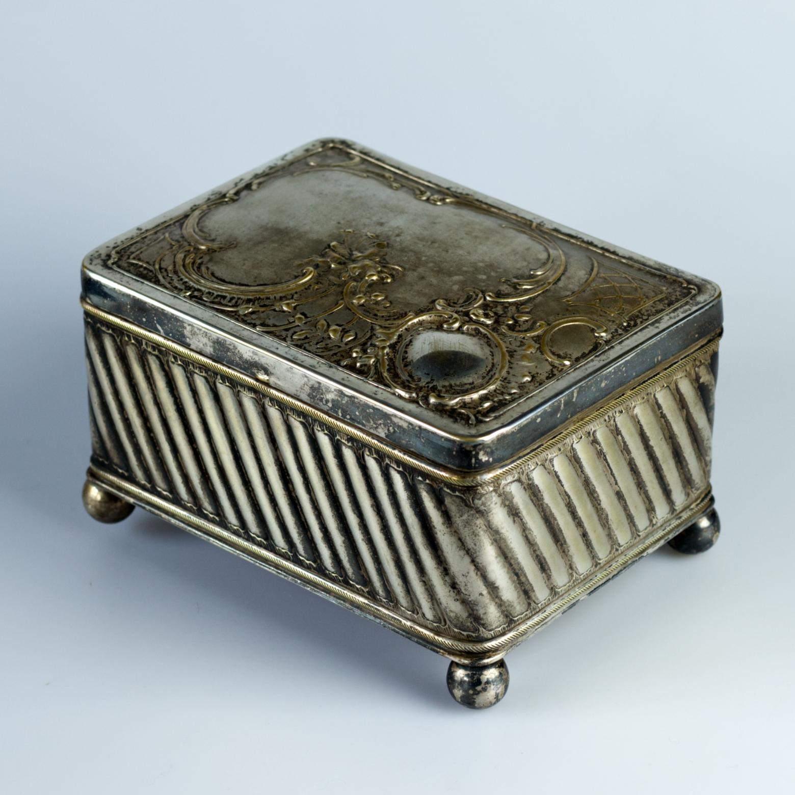 Scatola per sigarette o sigari placcata in argento con riempimento in cedro, decorata con rilievi di pregio. Si erge su quattro gambe sferiche e risale probabilmente alla fine del XIX o alla prima metà del XX secolo. Sul lato inferiore, con la