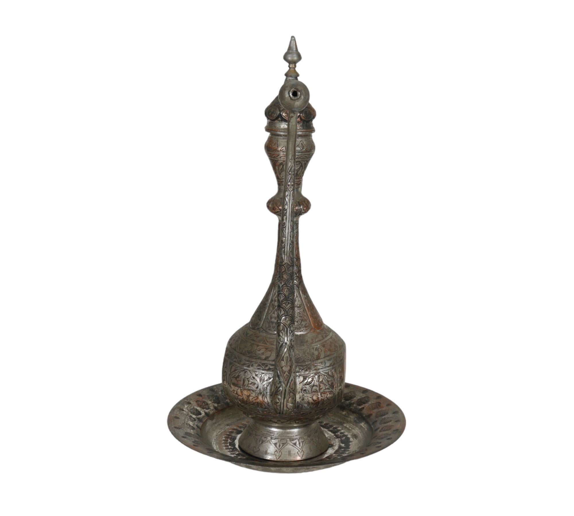 A large Middle Eastern silver plated copper ewer with matching serving plate. Tall with a long spout, neck and handle. Ornately carved throughout with a vine like motif.
