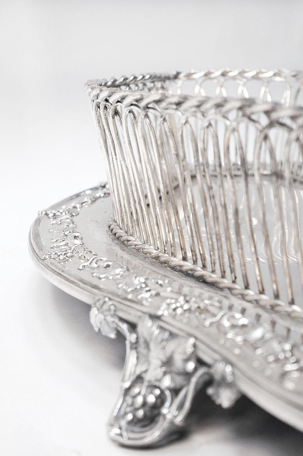 Silver plated metal centrepiece with a curve shaped plate adorned with a vine branches frieze on the edge, standing on four small curved legs with a similar decor. Plate topped by a sort of openwork wicker basket with arches shape edges braided up