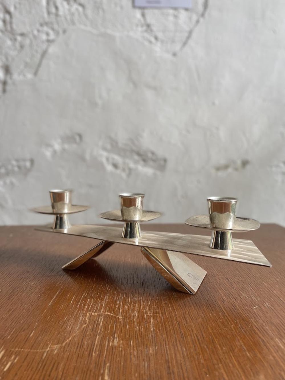 This modernist triple candle holder was likely produced in the 1960s during the height of Mid-Century Modern design. The piece features a distinctive geometric base with an angular, balanced form that exemplifies the clean lines and minimalist