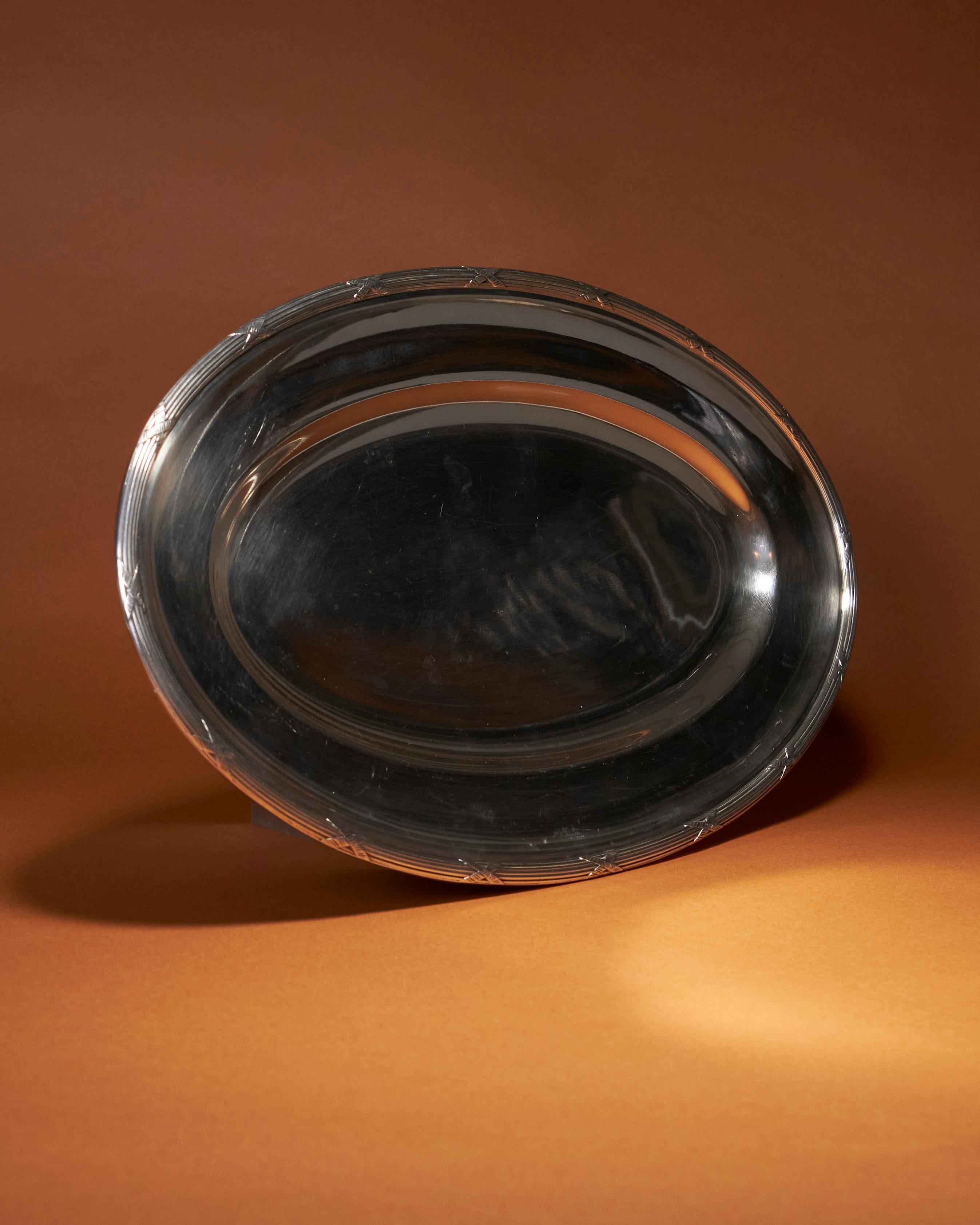 Plat de service ovale en métal argenté. Le bord du plateau présente de fines lignes, interrompues par de petites croix texturées. Ces détails ajoutent une subtile touche de raffinement et d'originalité à la pièce, renforçant son caractère élégant et
