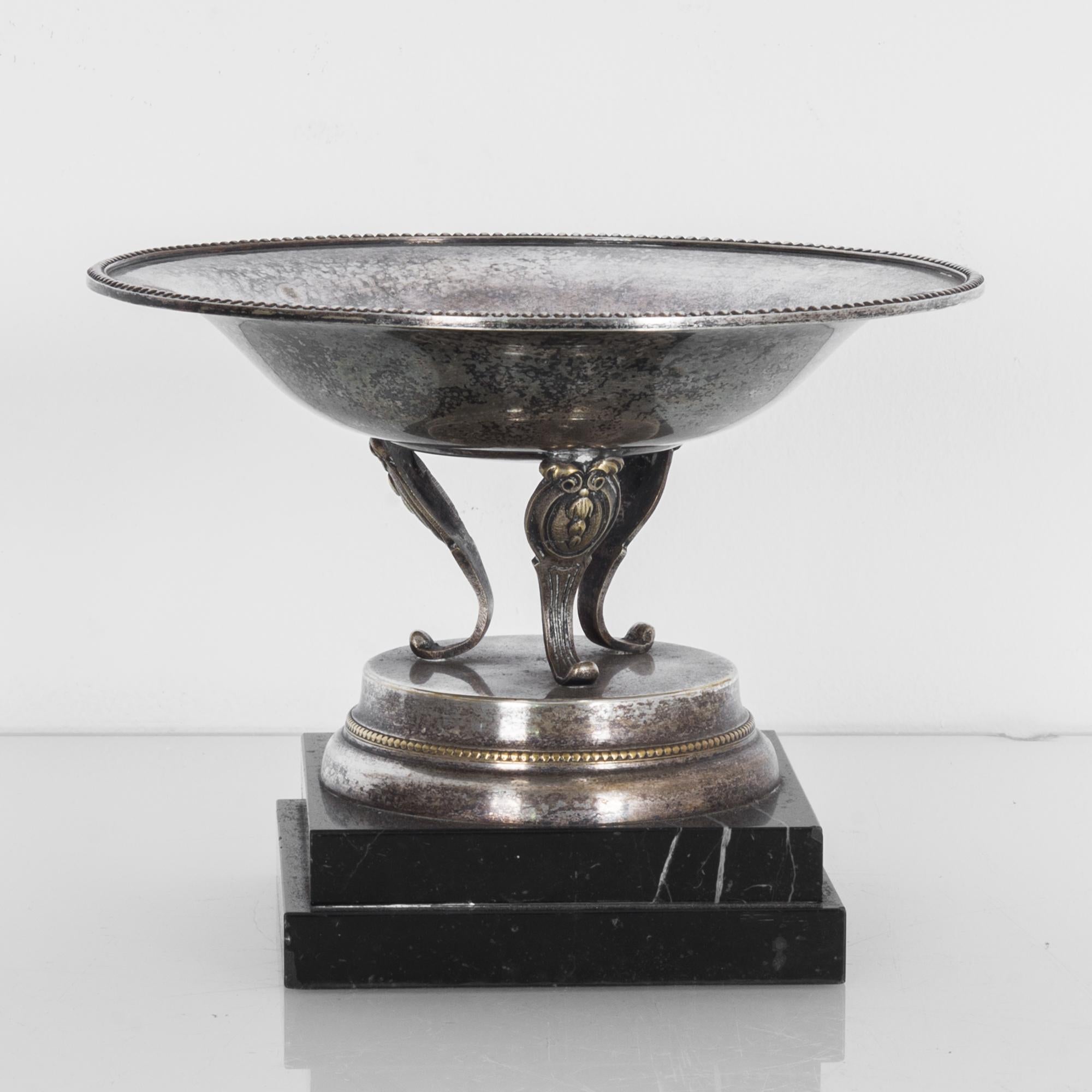 This silver plated trophy was made in Europe, circa 1900-1930. It features a wide, shallow cup with a decorative rim. The cabriole tripod legs display intricate carvings and rest on a circular pedestal. The trophy stands on a black, stepped marble