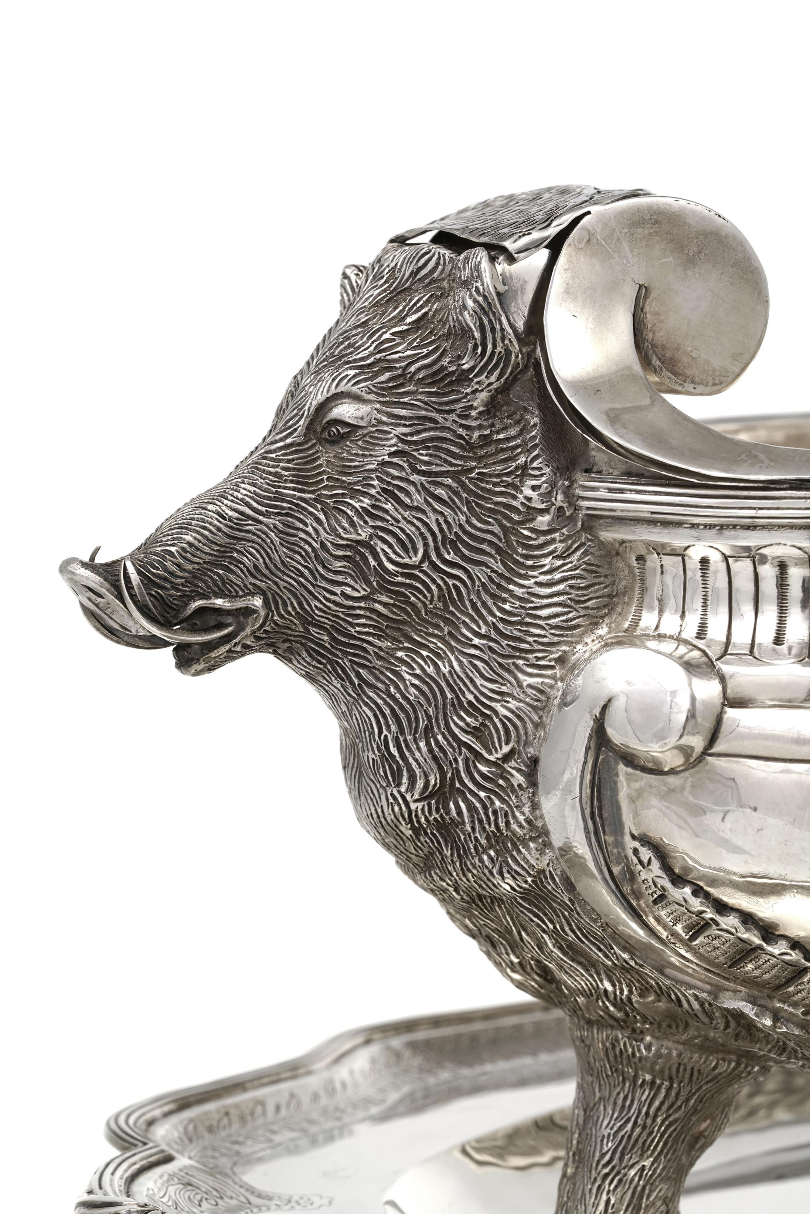 Elegante sopera bañada en plata con su forro extraíble y su soporte a juego, fabricada en el siglo XX. El cuerpo presenta un escudo central coronado en altorrelieve, enmarcado por volutas finamente esculpidas. Las asas de cabeza de jabalí añaden un