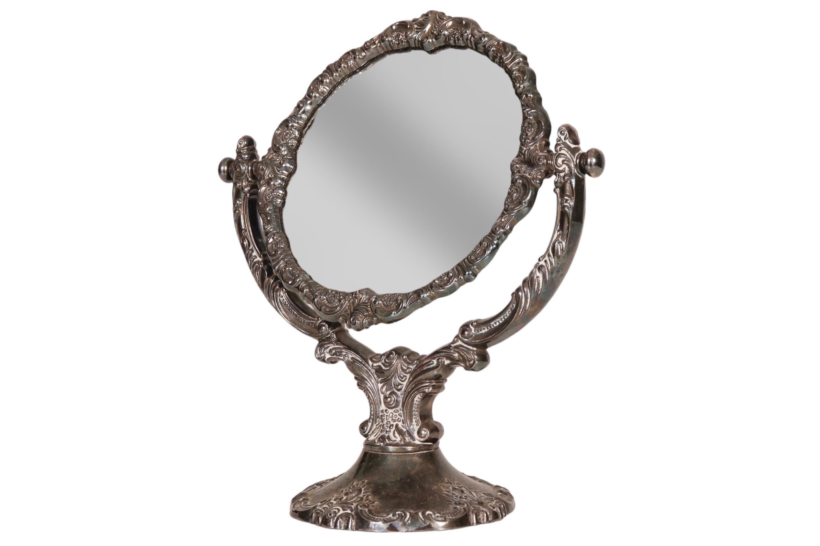 A 1940’s silver plated Baroque style vanity mirror made by Wallace. A round mirror is set in an elaborate adjustable frame, cast with c scrolls, feathers and beading. The back is finished with blue velvet.