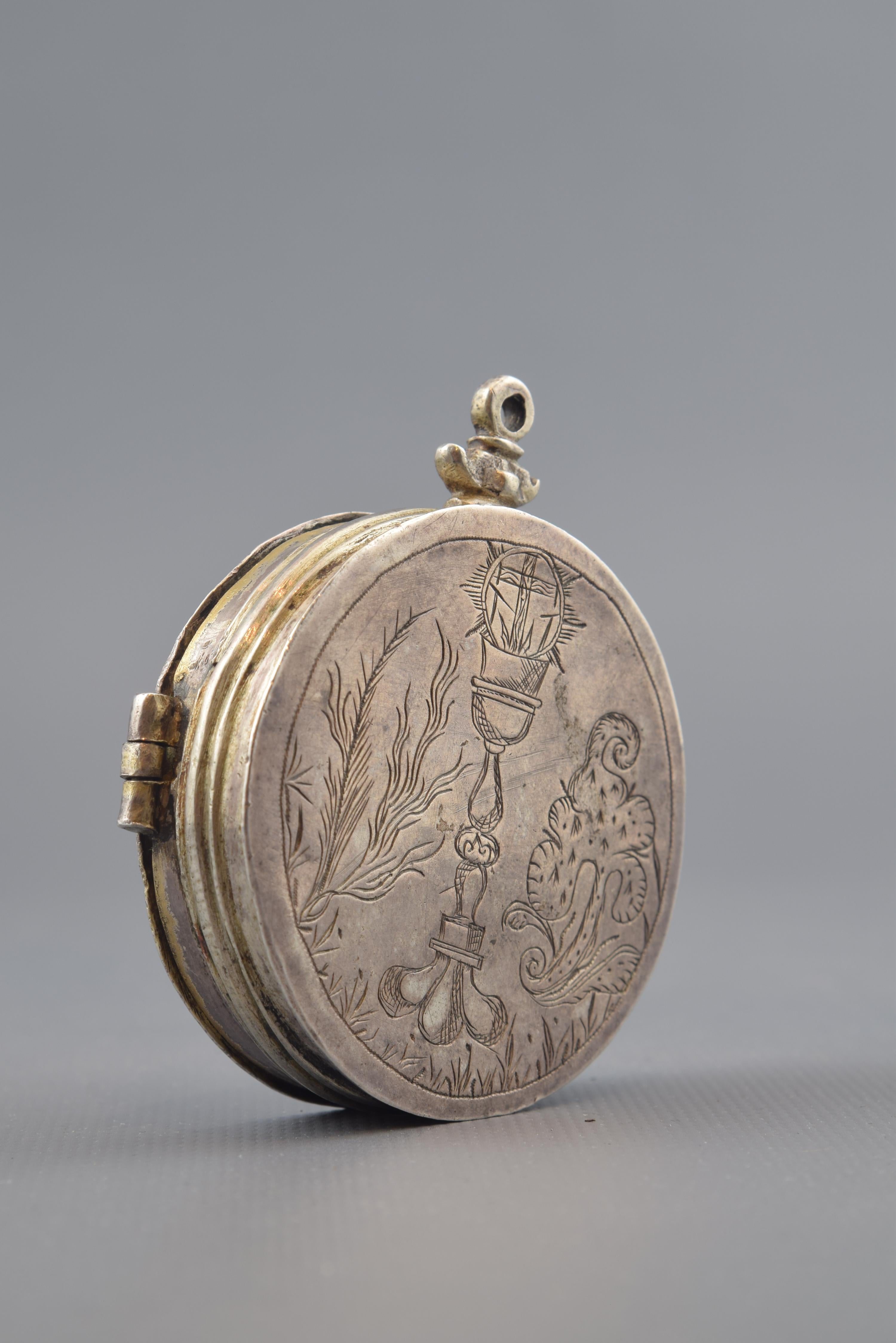 Silver box with a circular shape and a ring on the top (on an element reminiscent of a sculpture base) decorated on the side with smooth moldings and elements engraved on both sides of the piece. The lid is attached to the body by means of hinges