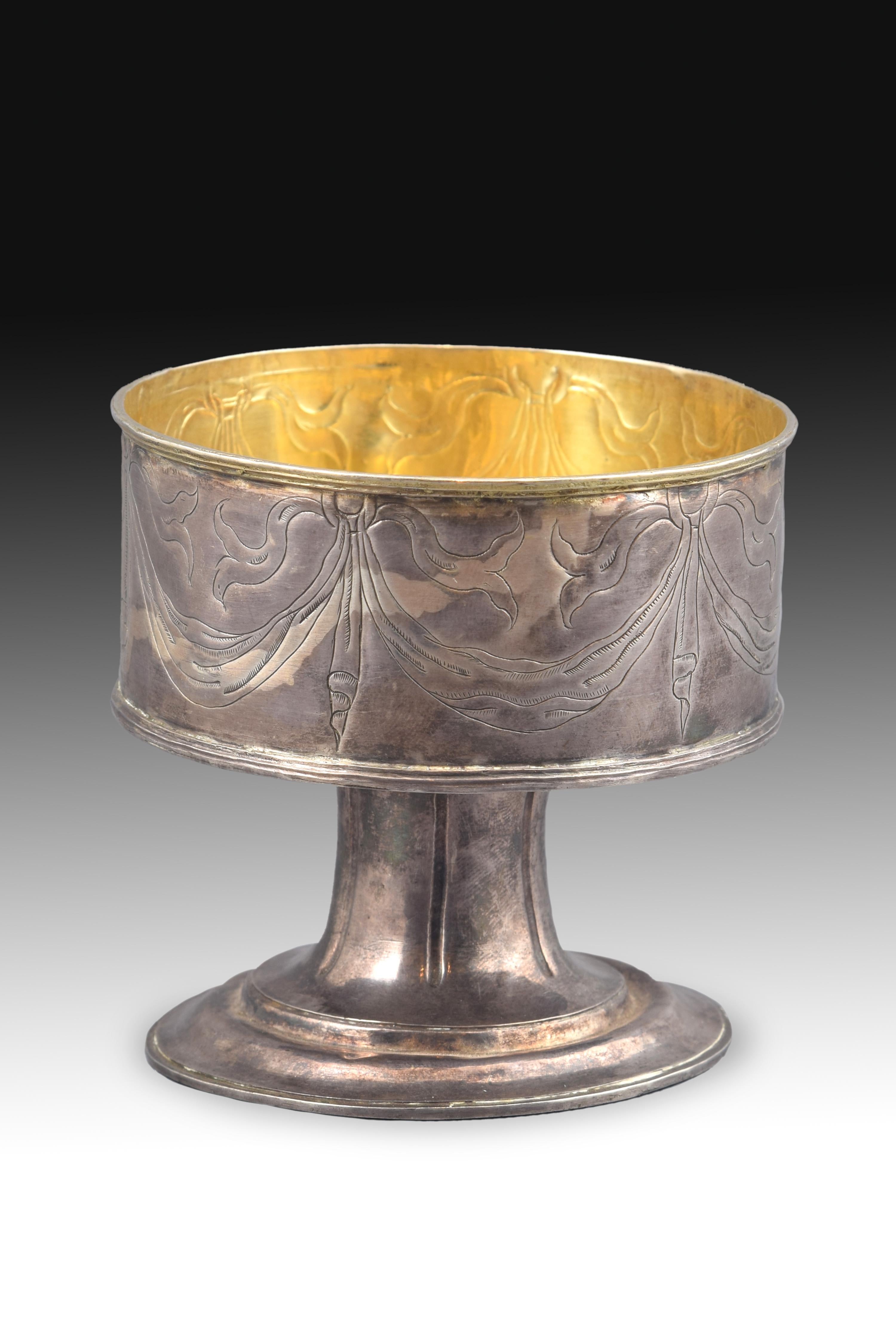 Piece made in silver (with the interior gilded) that shows a circular base elevated in two levels by smooth moldings and a grooved foot that leads into the container area, which is decorated on the outside with ribbons that hang from rings (this
