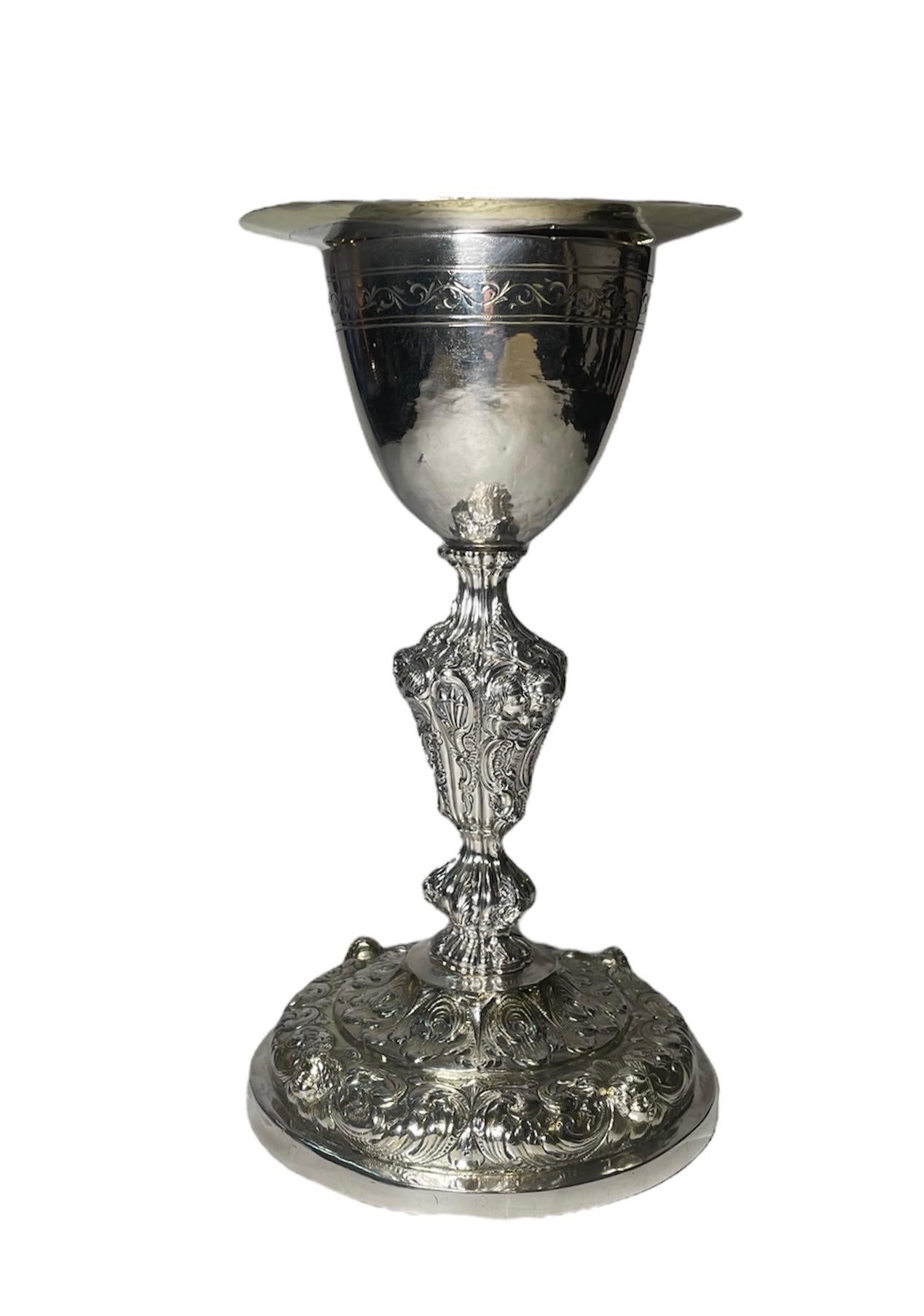 This is a Silver Chalice and Round Patten. It depicts a concave shaped cup adorned with an engraved garland of scrolls branches of leaves at the upper border. It is supported by a repousse stem decorated with cherubs’ faces, rocailles and scrolls of