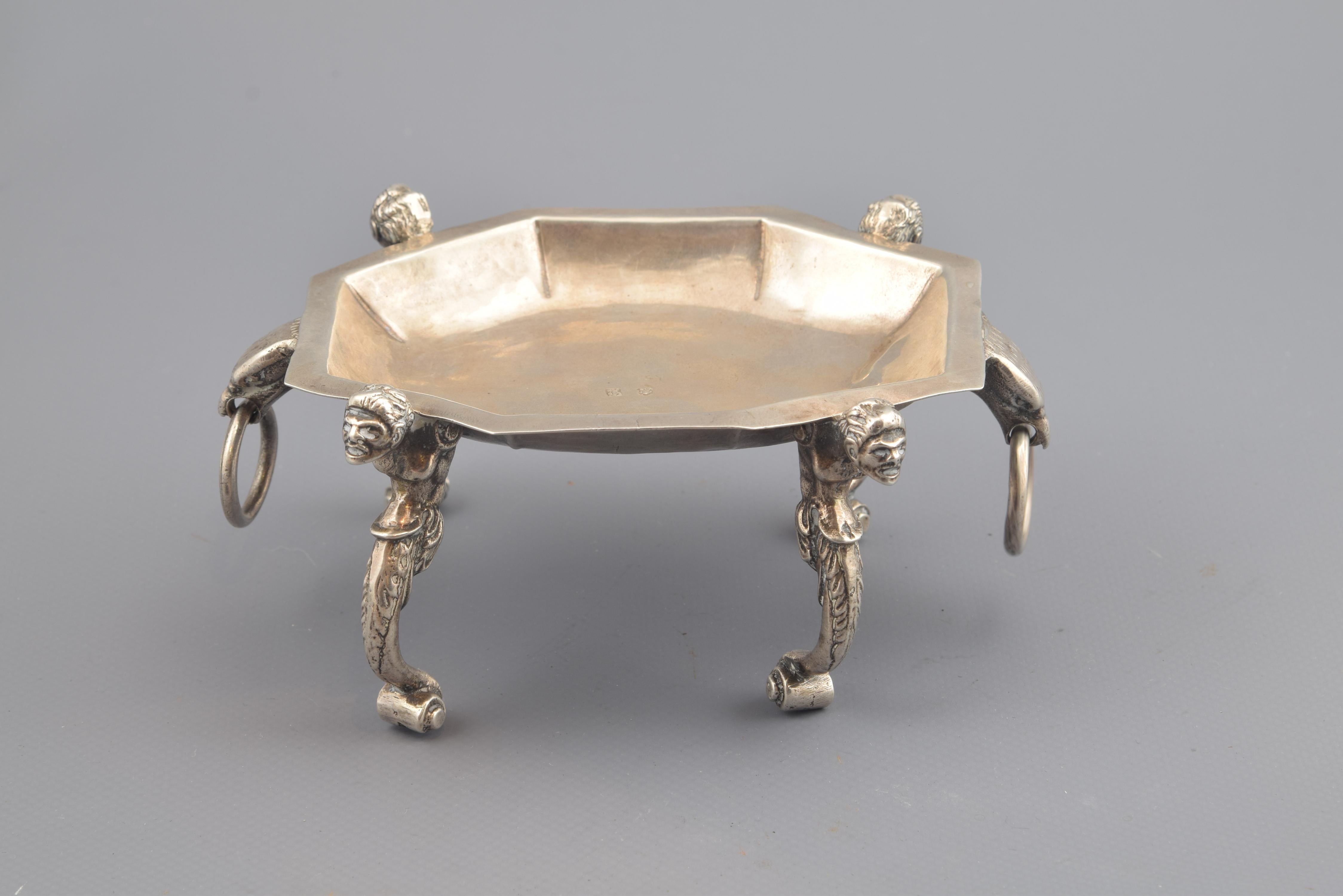 Specie or silver salt cellar in its color composed of a tray not very deep polygonal, raised and enhanced by four legs and also has two protomes of animals holding rings (eagles or lions). The legs have a lower scroll finish, which gives way to a