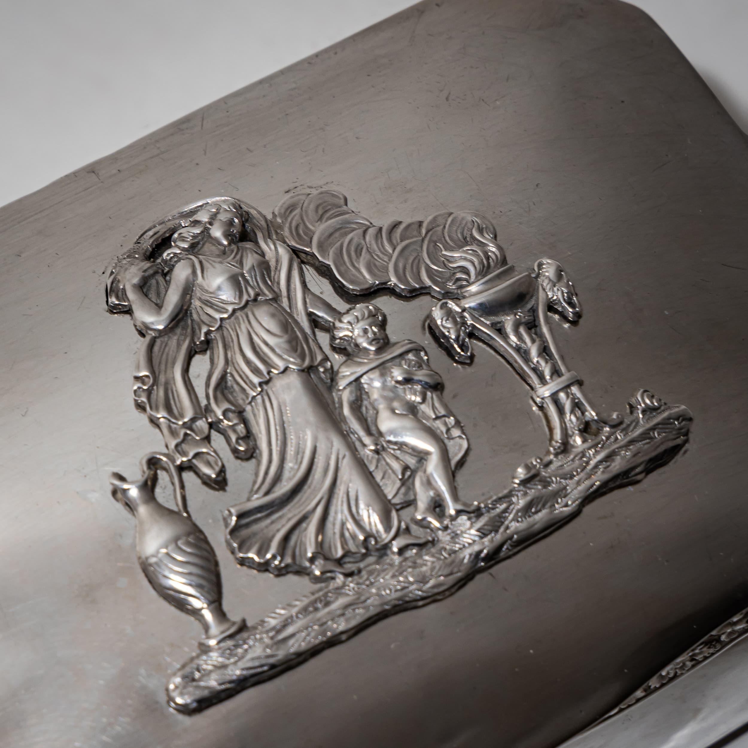 Azucarero de plata, pequeño y finamente labrado, apoyado sobre patas con cuerpo octogonal. La tapa presenta un friso decorativo de hojas de vid alrededor del borde y un detallado aplique en relieve que representa a Venus y Cupido junto a un ánfora y