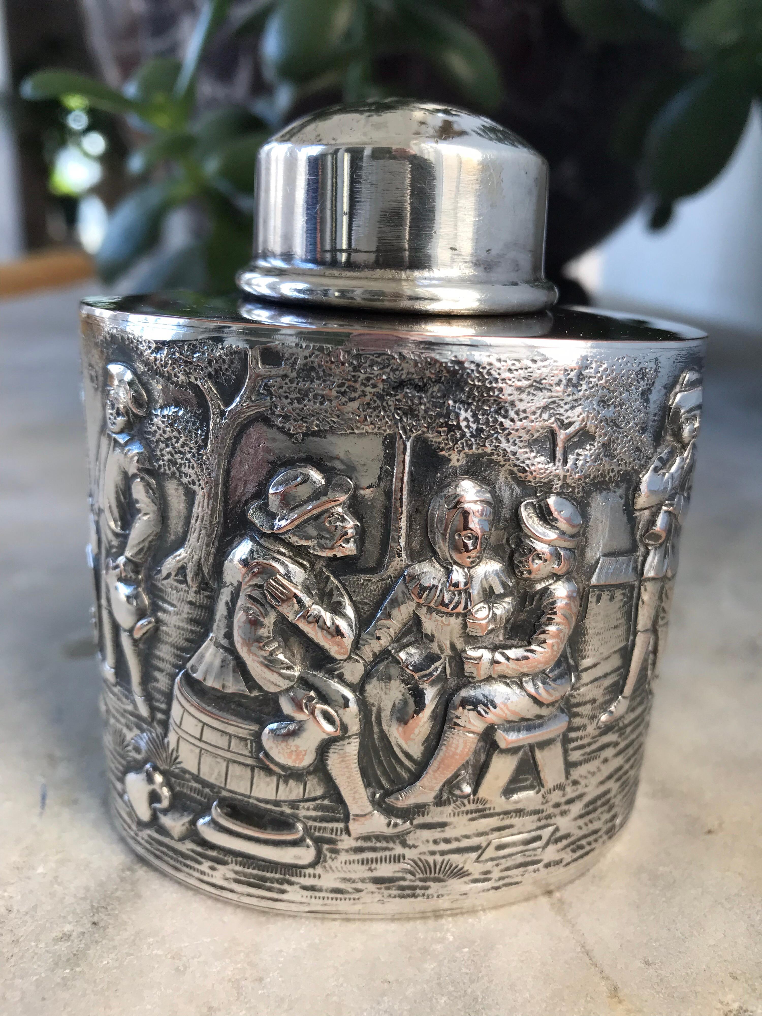 Antique silver plated tea caddy with high reliefs of a village scene surrounding the entire body of the tea caddy, illustrating musicians, dancers, taverner, a group socializing and drinking, and a very charming dog looking on. This quality caddy