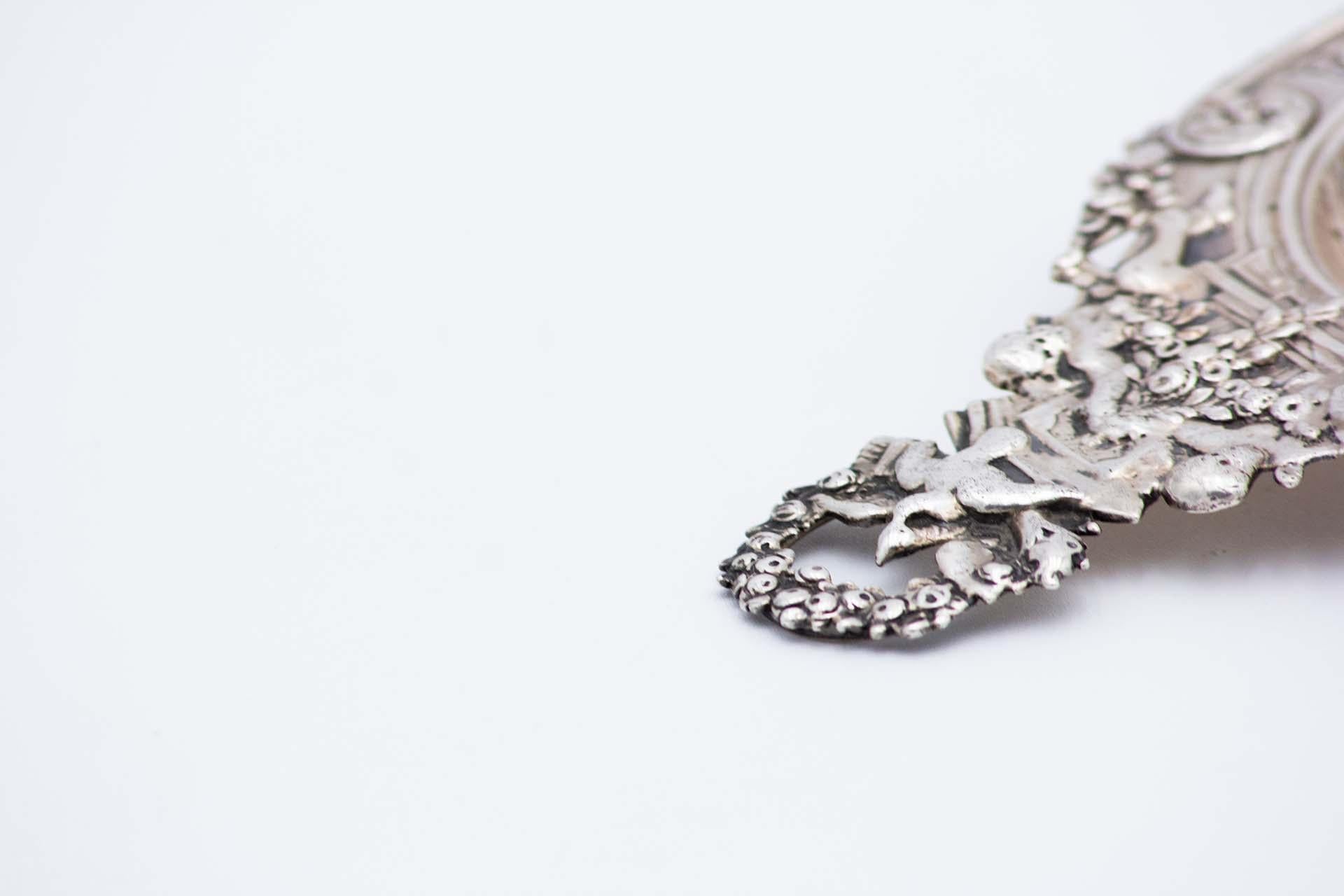 Description : Passoire à thé en argent avec des Puttis en relief, des nœuds, des couronnes et des palmiers abondamment définis. Métal : Argent. Origine : Suède. Marque : Trevo. Poids : 81,75 grammes. Dimensions : 15,3 cm x 2,3 cm : 15,3cmx2,3cm.