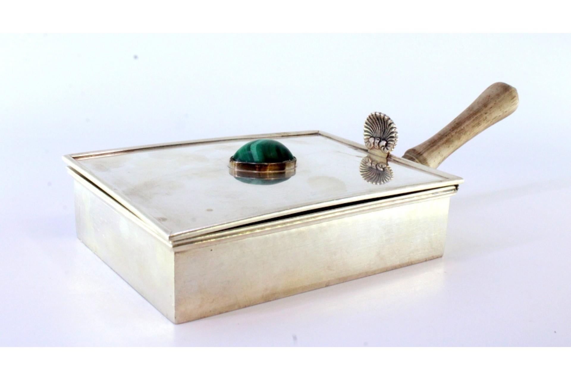 Description: Silver cigarette holder with wooden handle and interior, shell appliqué on the lid and fake stone in the center. Metal: Silver. Origin: Portuguese. Mark: Standing Eagle I Title. Weight: 433.17 grams. Dimensions: 21cm x 10.2cm. Content: