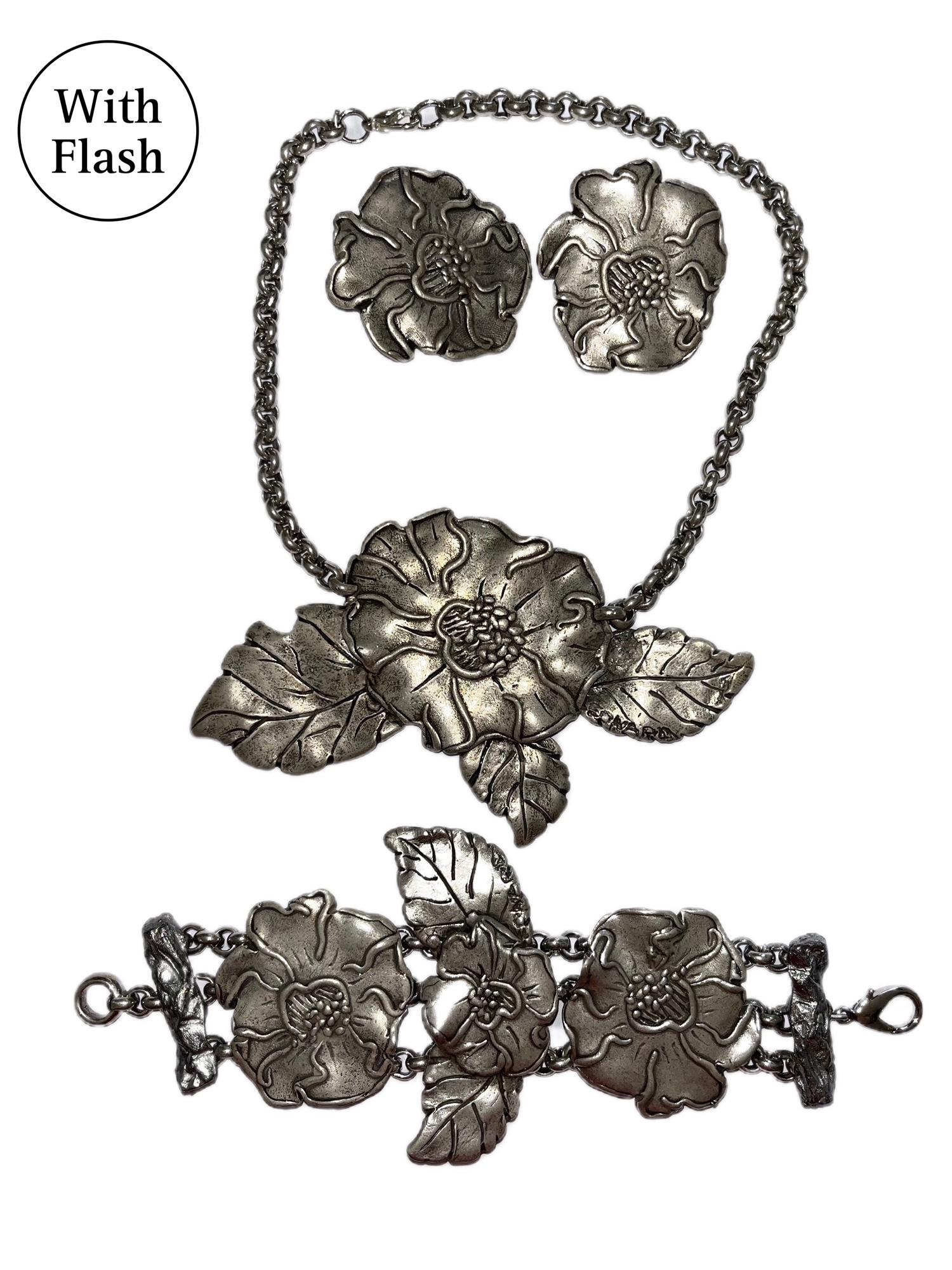 This three-piece set is composed of matching silver-toned clip-on earrings, a chain bracelet and a chain necklace. The three pieces of jewellery by Leonard of Paris have as the centrepiece wild roses. 
Each clip-on earring is composed of a wild