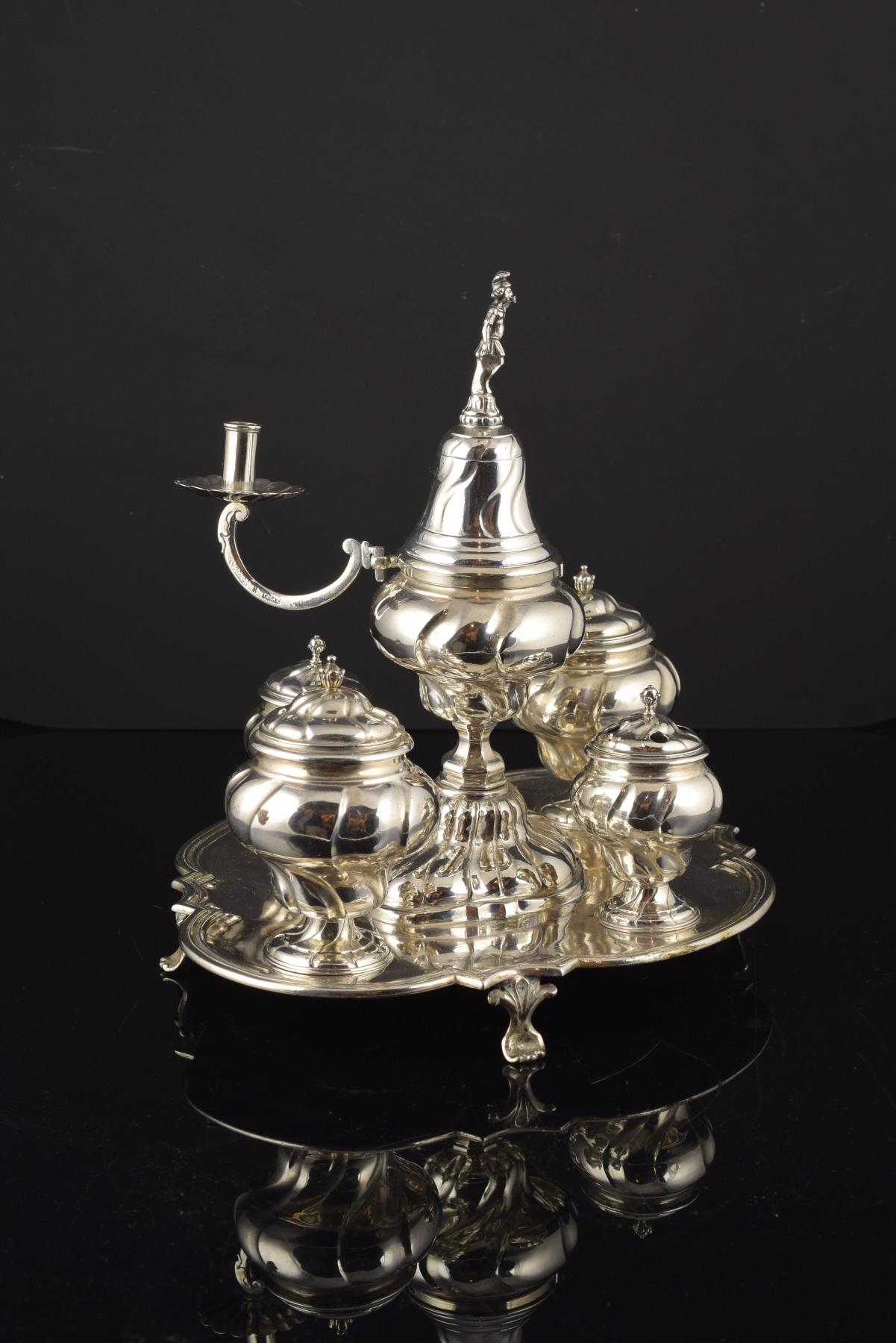 With hallmarks.
The base tray has a square shape with mixtilinear profiles and rests on legs with vegetal motifs. The four containers present helical gallonized feet, and pearl flower finials on the covers. A candlestick emerges another container,