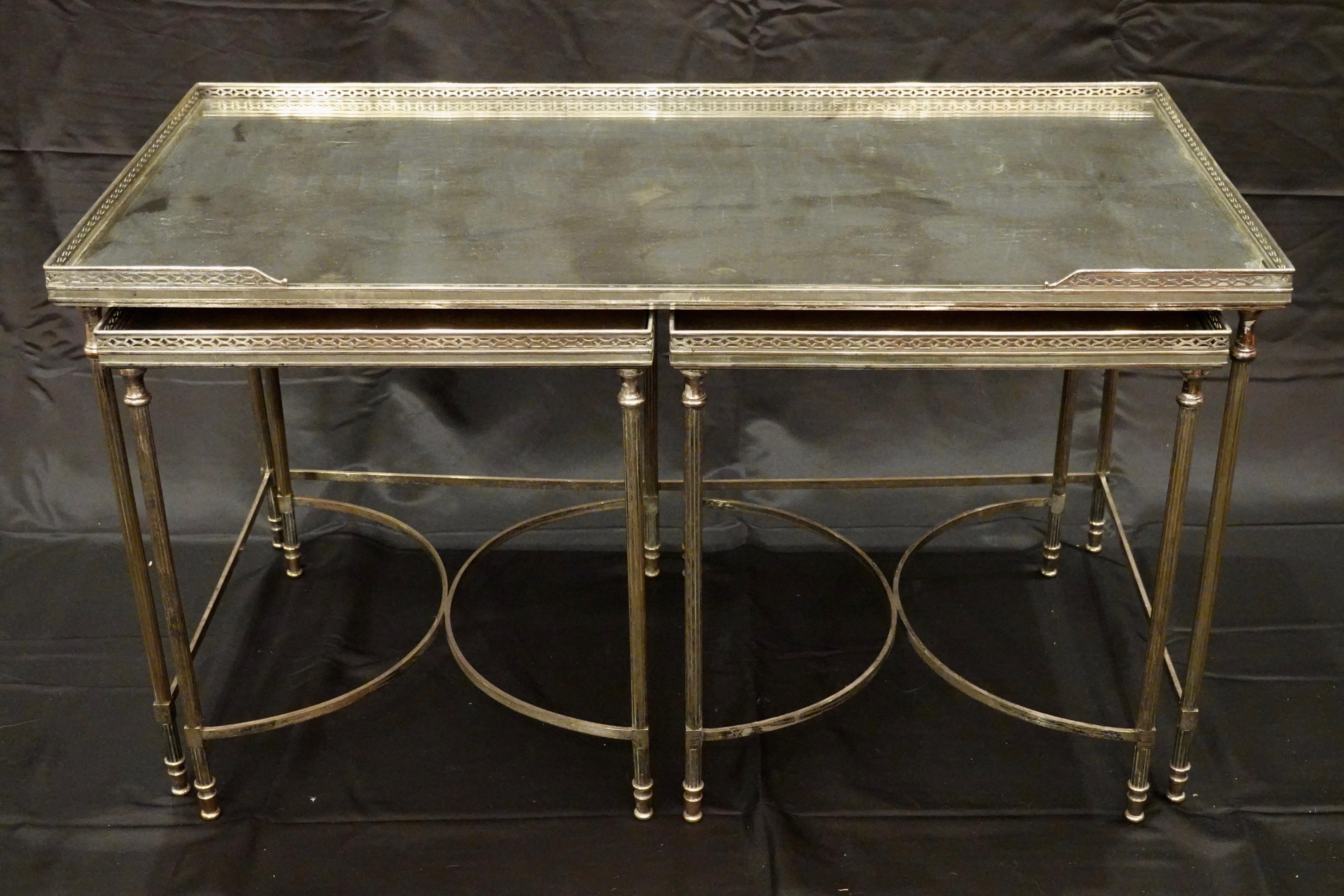 A set of three silvered-bronze tables comprising a coffee table and two matching side tables that nest under the coffee table. The tops are églomisé silver with tones of gold and grey veining. The tables feature an elegant neoclassical design with