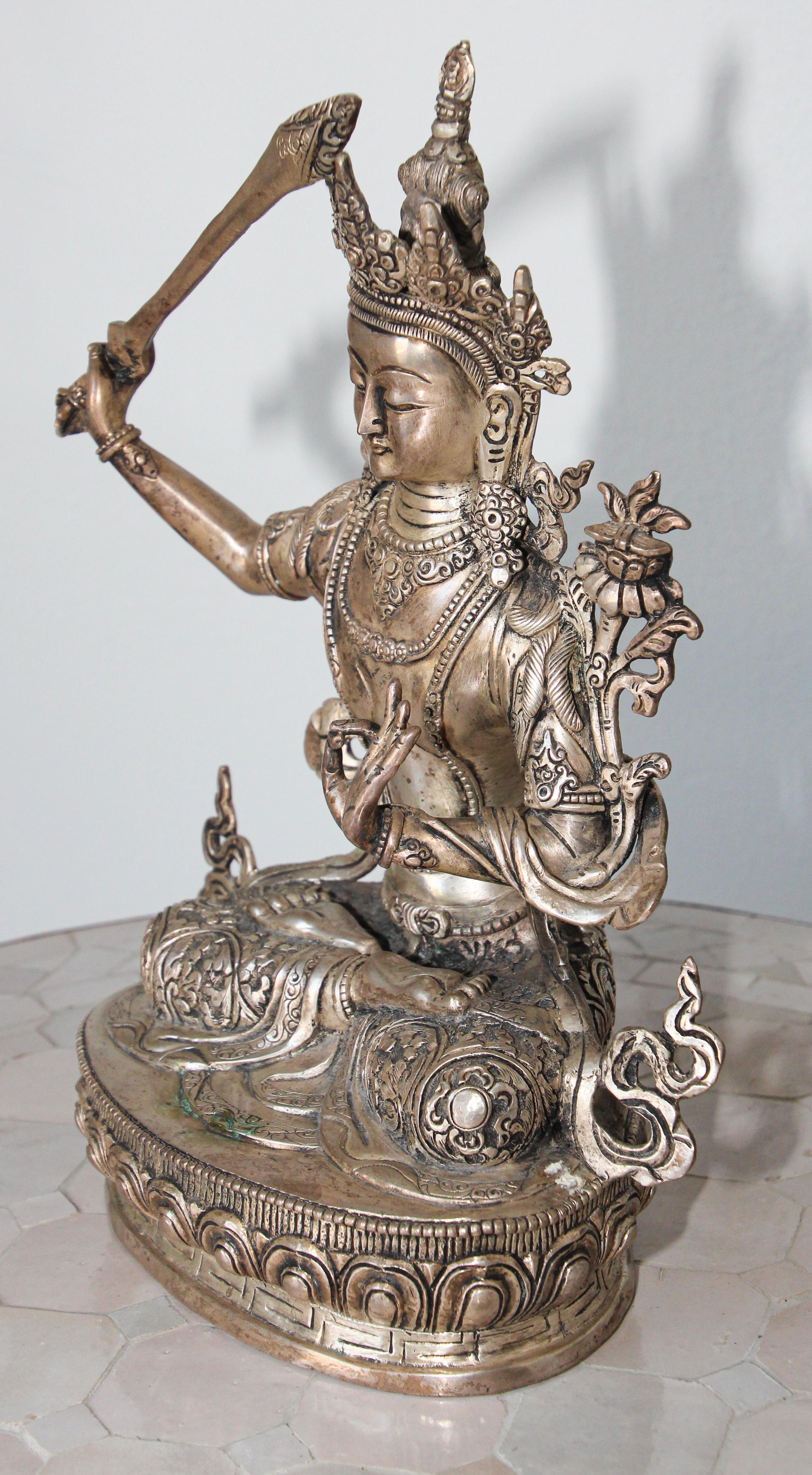 A silvered figure of Manjusri seated in dhyanasana on a Pala style Indian base, wearing an ornate crown and wearing body jewelry,
Manjushri holds the sword of wisdom in his right hand and above his left shoulder is a lotus flower upon which rests a