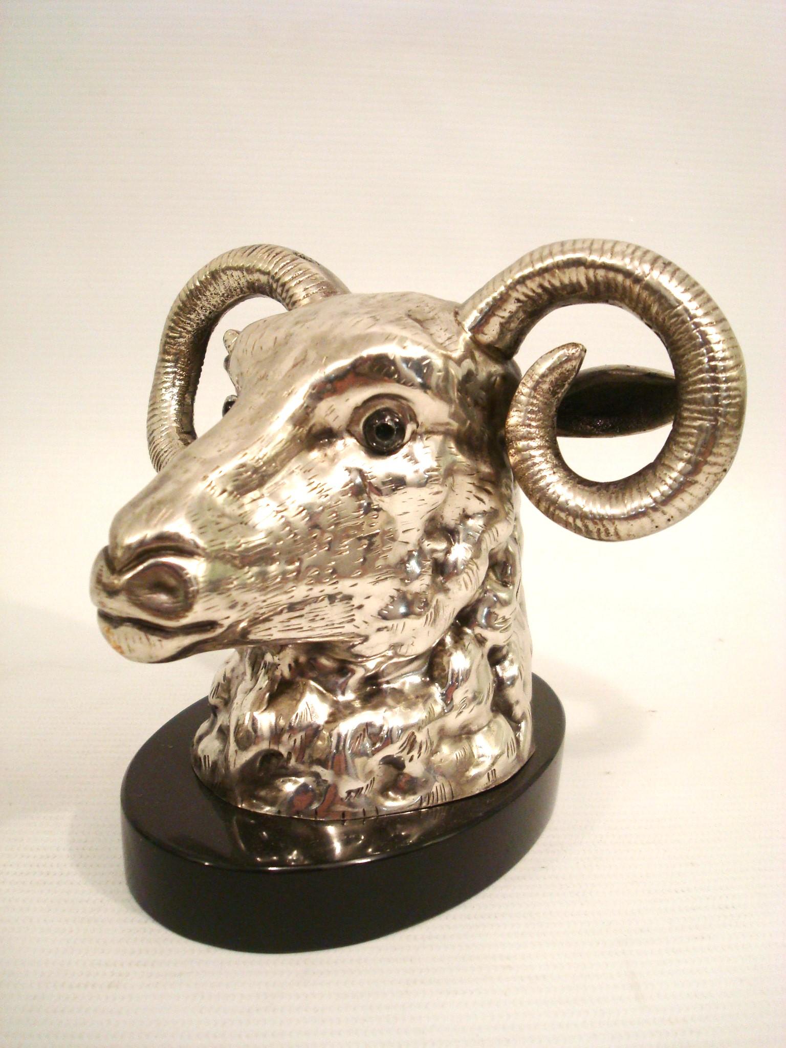 Sculpture en métal argenté représentant une tête de bélier avec des traits détaillés et des yeux en verre, notamment des cornes incurvées et une fourrure texturée. La sculpture est montée sur un socle en marbre noir. La qualité de l'exécution et les