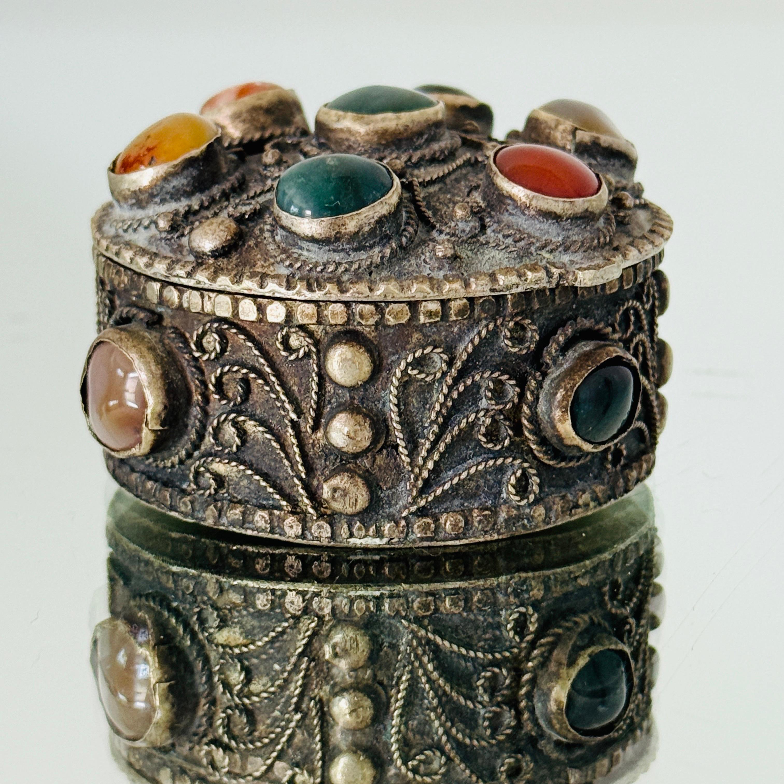Handcrafted silvered metal trinket box with insets of semi-precious stones in hues of red jasper, green malachite, yellow citrine, and red carnelian agate. The round box has a series of hammered beads and hand carved or engraved scroll motifs. The