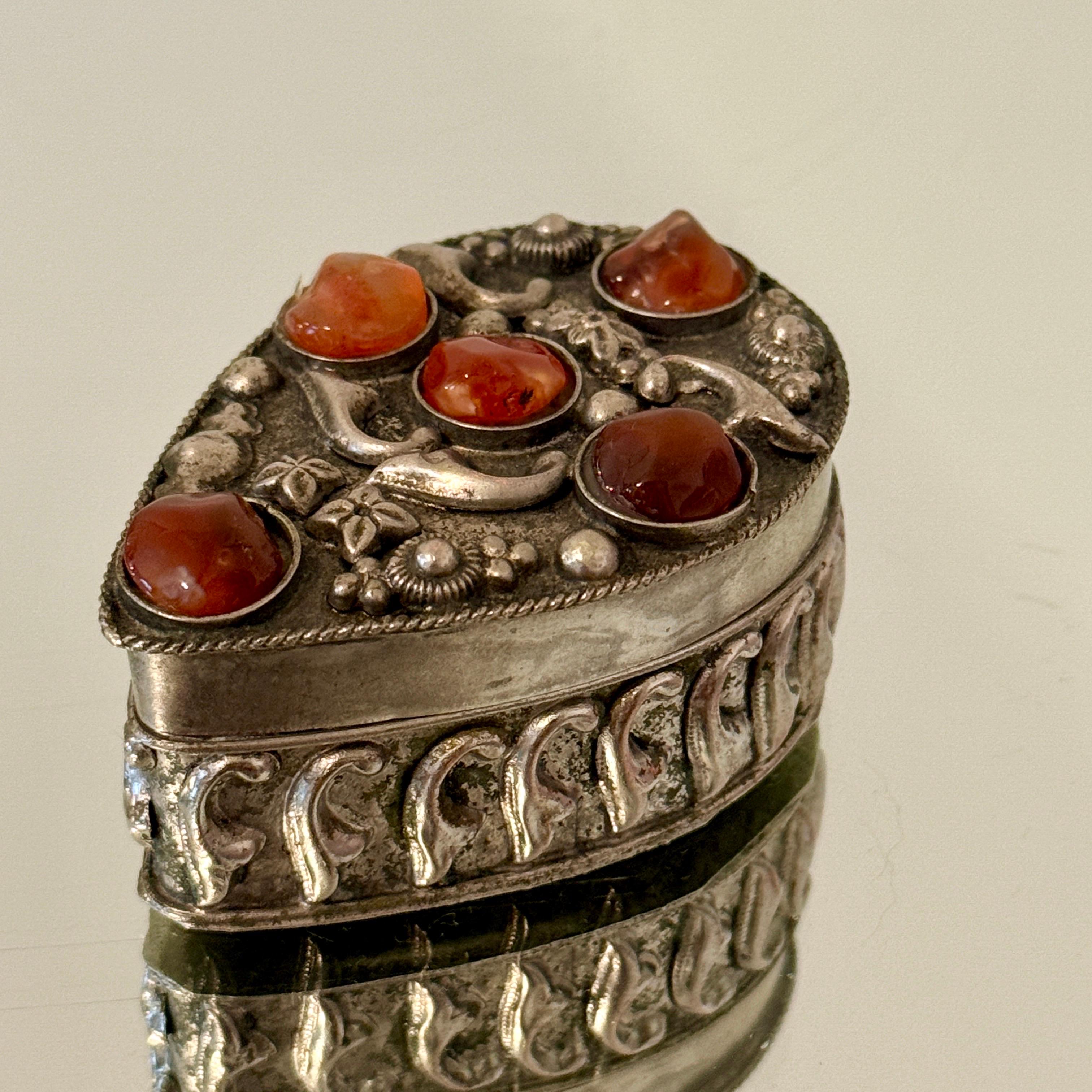 Handcrafted silvered metal trinket box with insets of semi-precious stones in hues of red. The box has a series of hand carved and incised scrolls and floral motifs, and has a teardrop form. The lid opens to reveal a velvet interior in dark purple.