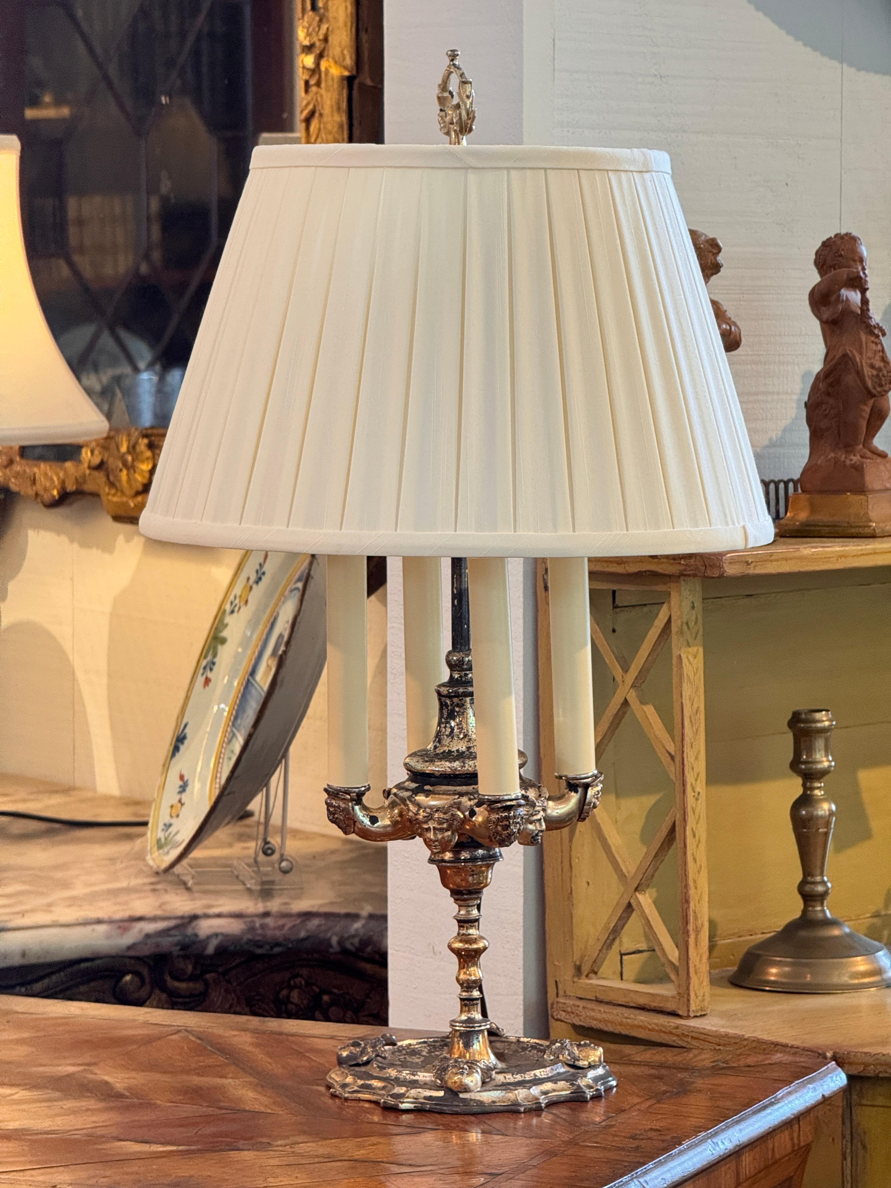A silverplate candelabra-form lamp with four faux candle lights set on an ornate classical base. The design incorporates masks and scrollwork in high relief, with a timeworn finish showing attractive wear to the plating. Topped with a pleated cream