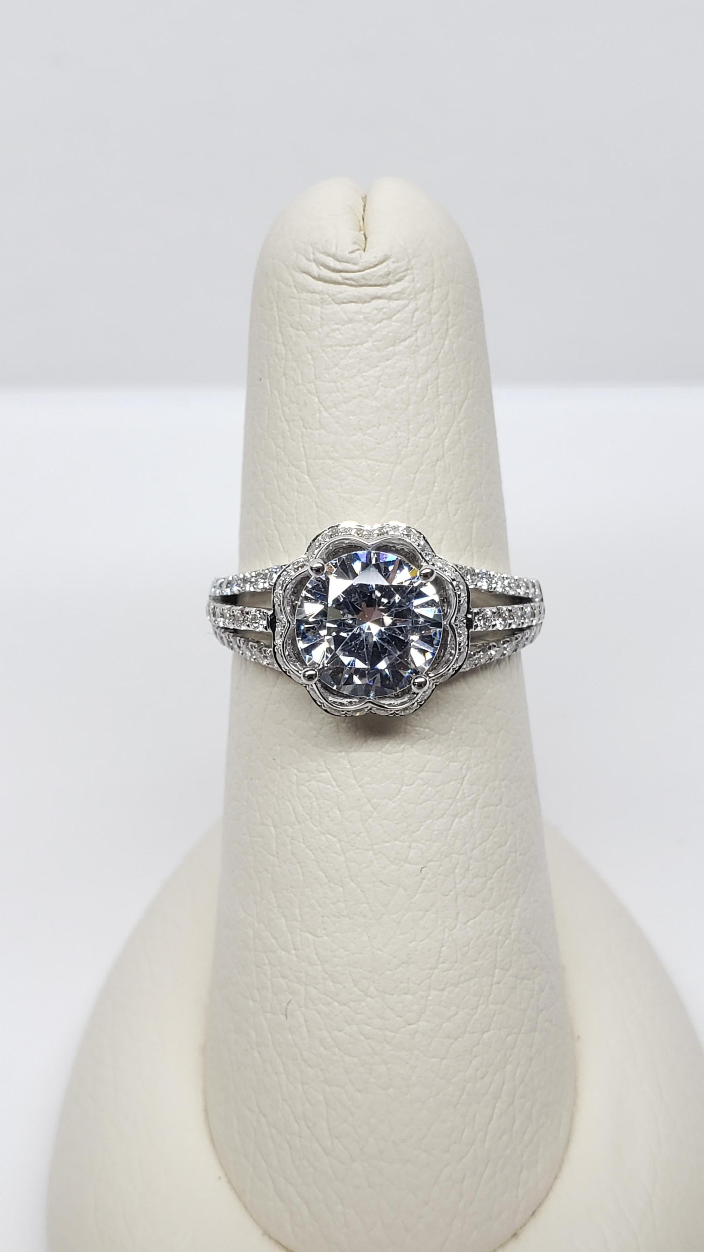 This semi mount was designed by Simon G. and is made from 18k white gold. It features a flower shaped diamond halo, a double split shank with pave diamonds along the upper half. The gallery has some delicate scroll work, and the center stone is set