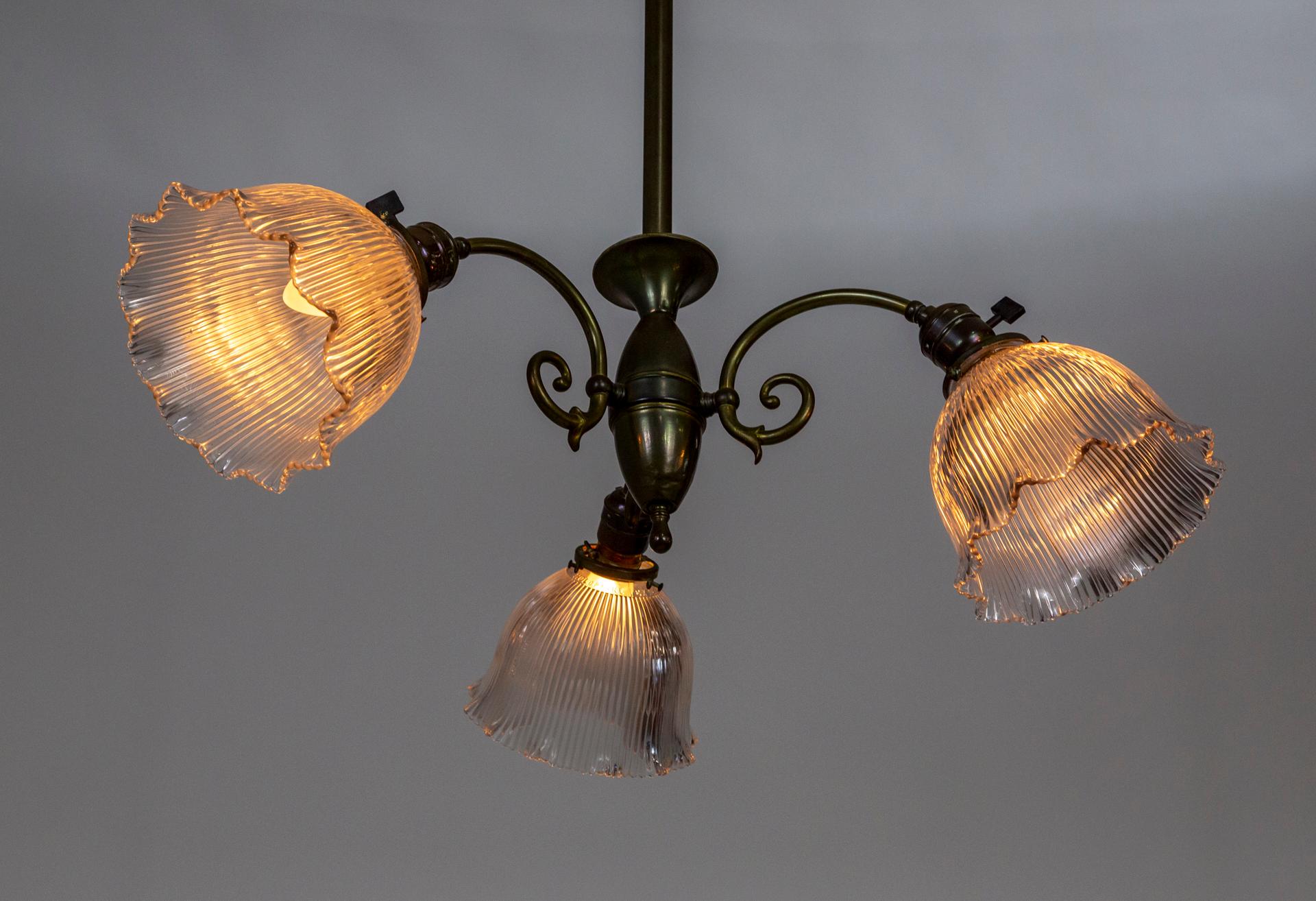 A rich, deep patinated brass chandelier and three curled arms with down lights. A simple, elegant design with ruffled bell shape, holophane glass shades. With switches on the sockets. Newly wired. Shades are marked 
