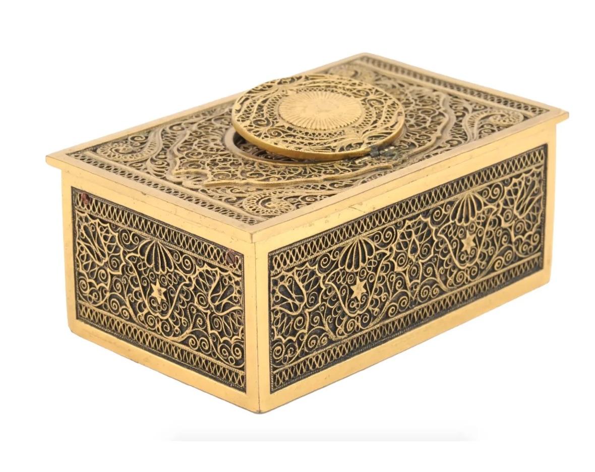 A mid-century gilt brass wind-up music box. The piece is decorated with ornamental filigree. A lid on top of the box reveals a singing parrot. Hallmarks Made in Germany, Ken-D, KG are on the bottom. Karl Griesbaum KG was a German manufacture that