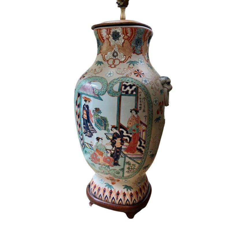 A single chinoiserie style table lamp with a figural scene on two sides surrounded by intricate colorful designs.  Lamp sits on a wood base and includes a pleated shade with trim. Lamp depicts  Asian women in traditional dress surrounded by trees